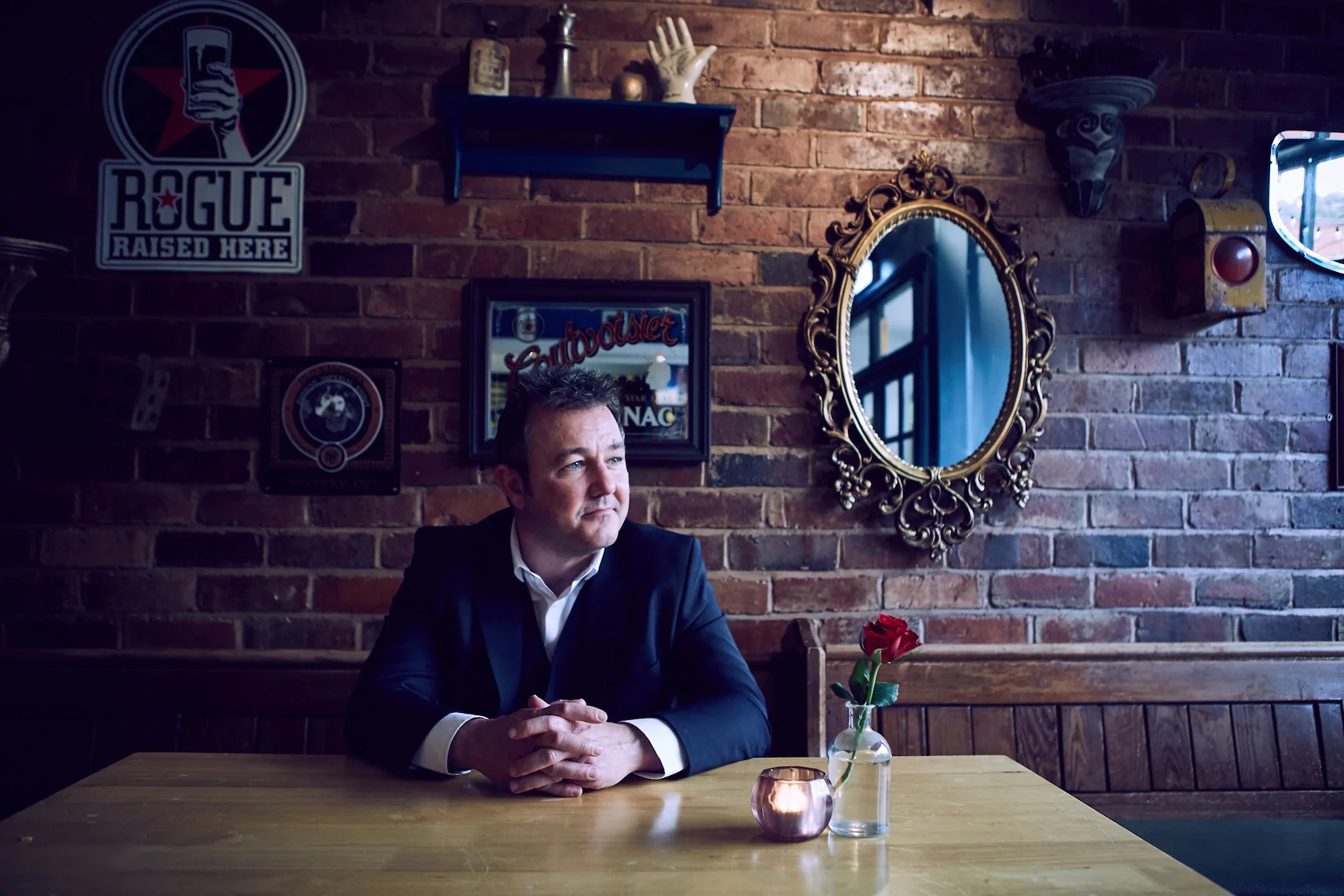 A man in a suit sitting at a wooden table in a cozy, dimly-lit cafe or restaurant with brick walls. Behind him are decorative signs, mirrors, and shelves with decorative objects. A single red rose in a glass vase and a small candle on the table add t
