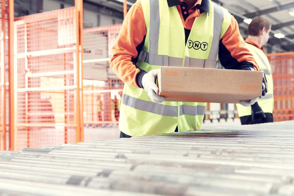 Worker in high-visibility vest and gloves, holding a cardboard box in a warehouse or distribution center.