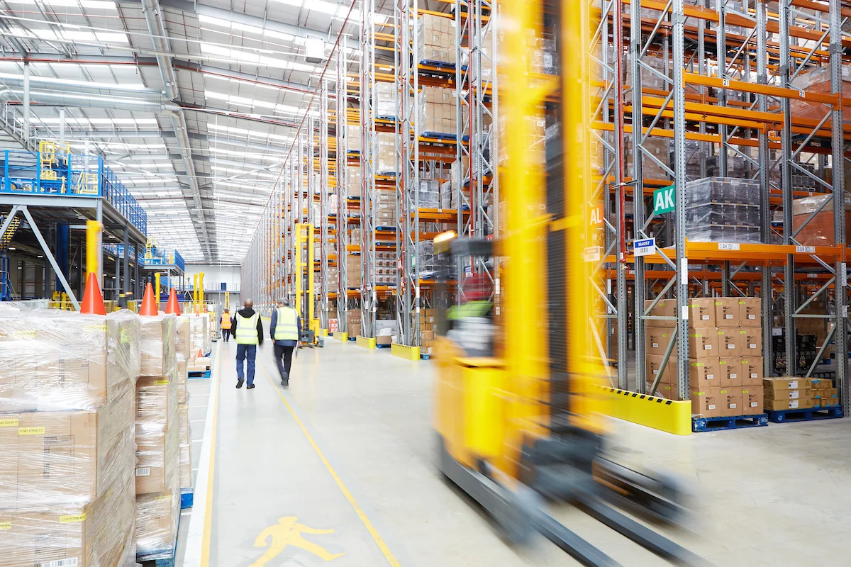 Inside a large warehouse with high ceilings, shelves filled with boxes and pallets, workers in safety vests, and a yellow forklift in motion.