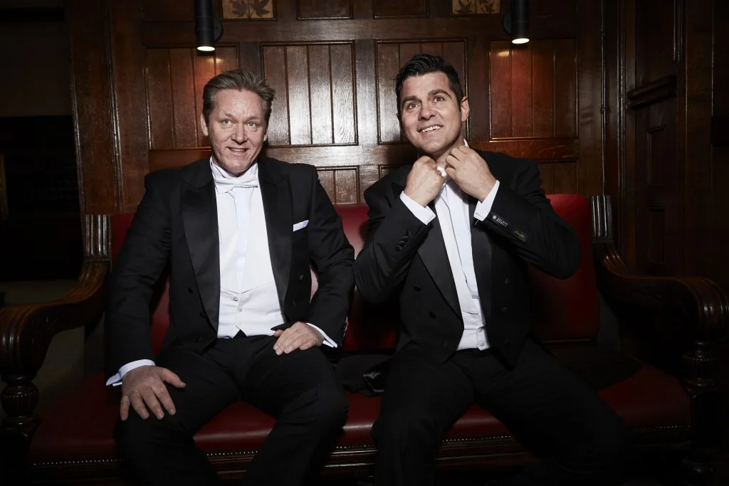 Two men in tuxedos sitting on a red leather bench inside a wooden-paneled room, smiling at the camera.