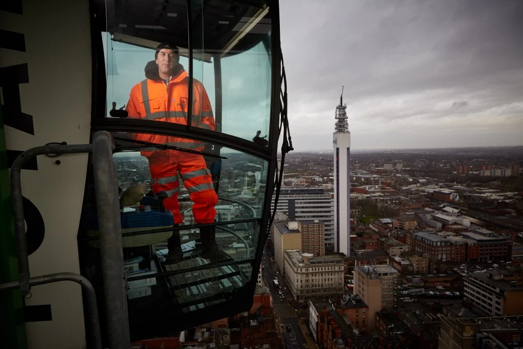 A worker in an orange safety jacket and pants standing inside the observation cabin of a tall building, with a cityscape and a tall communication tower in the background.