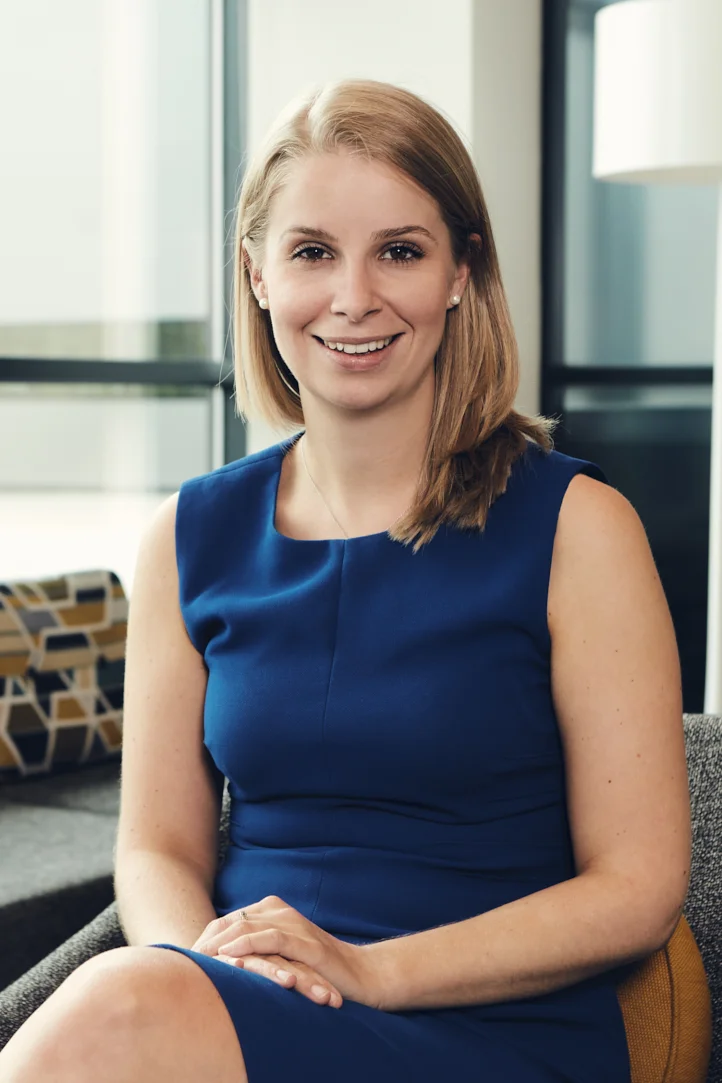 A smiling woman with shoulder-length blonde hair wearing a sleeveless blue dress, sitting on a gray chair in a modern office with large windows and a patterned cushion in the background.