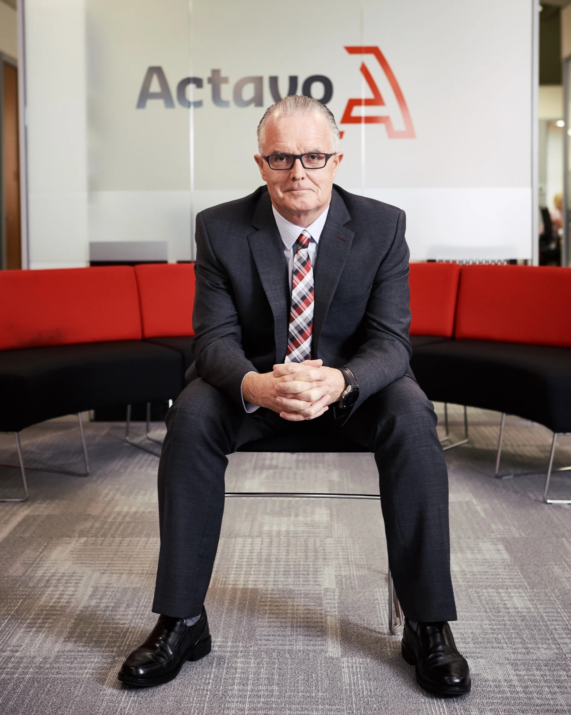 A man in a business suit sitting on a black and red curved bench in an office lobby, with a logo that says 'Actavo' behind him.