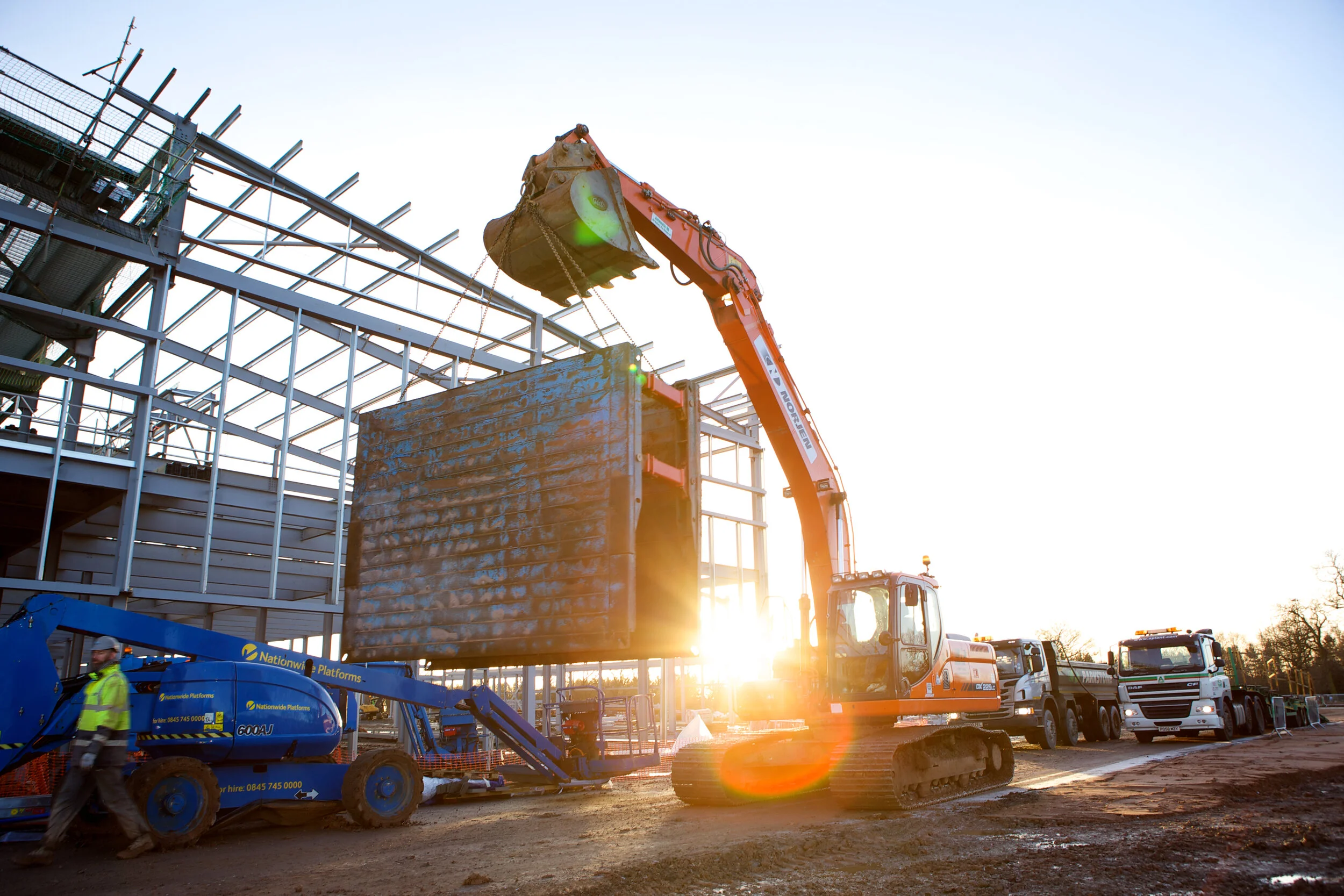 Construction site with an orange excavator lifting a large steel section, with workers and trucks visible in the background at sunset.