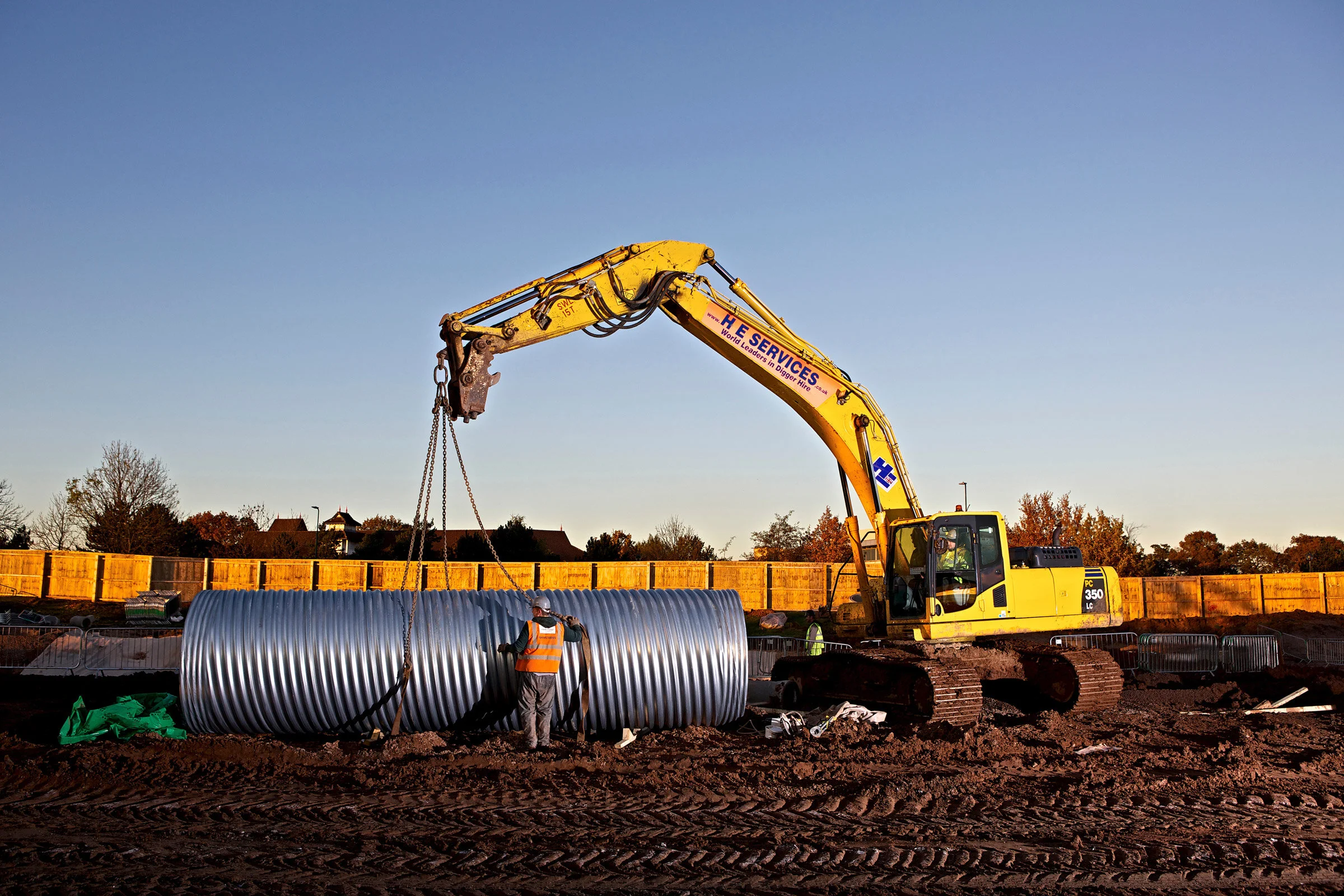 Construction site with a yellow excavator lifting large metal pipes, with workers inspecting the pipes and a wooden fence in the background under a clear sky.
