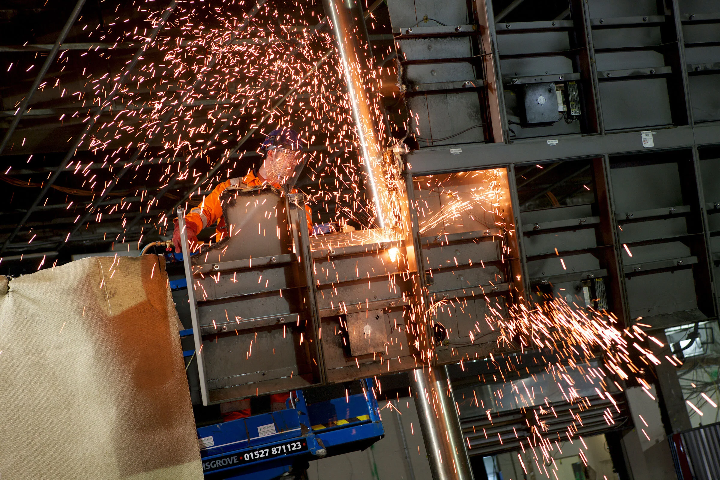 A worker in safety gear welds metal on a construction site, sparks flying from the welding process.
