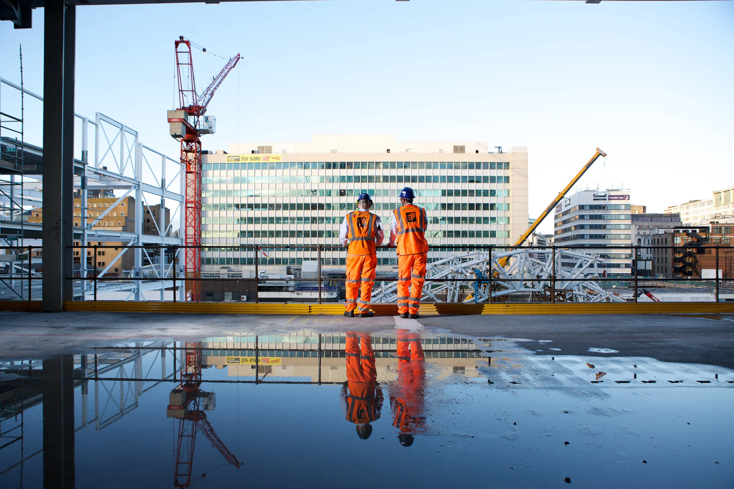 Two construction workers in orange safety uniforms and blue helmets standing on a construction site, looking at the cityscape with cranes and buildings in the background, reflected in a puddle on the ground.
