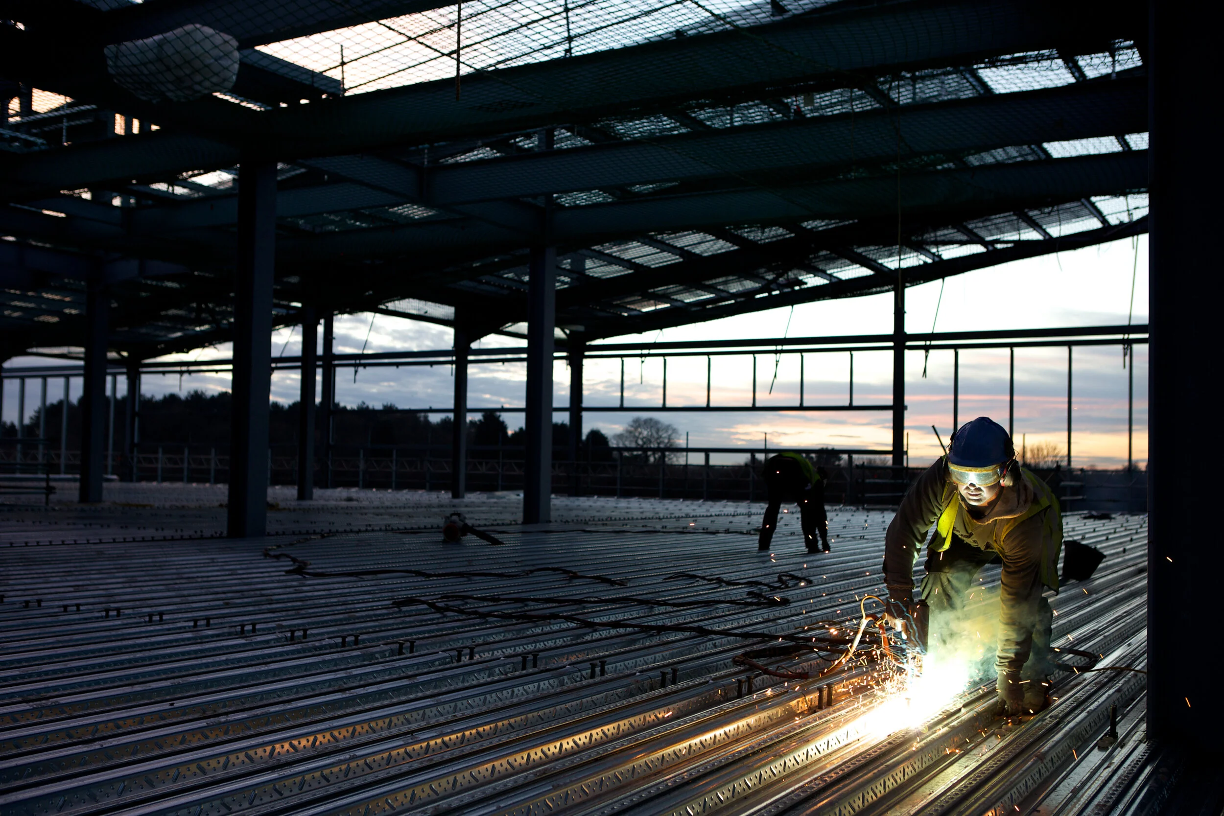 Two construction workers welding on a metal framework at sunset, with a partially built structure overhead.