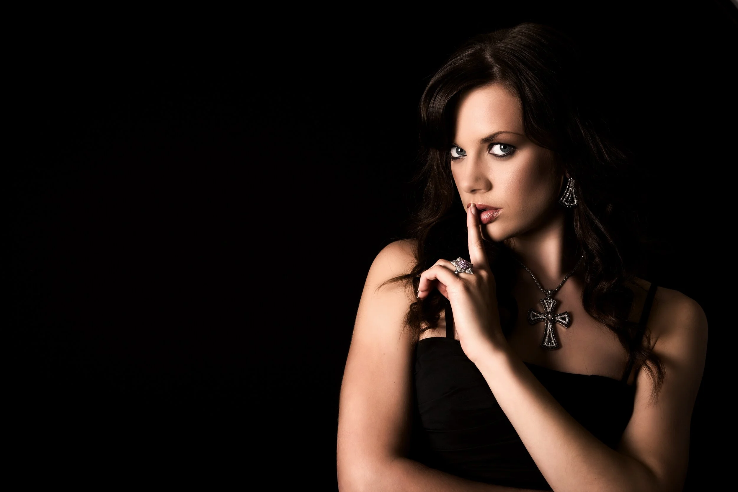 A woman with dark hair and blue eyes wearing goth-style jewelry, including a cross necklace, earrings, and rings, posing with a finger to her lips against a black background.