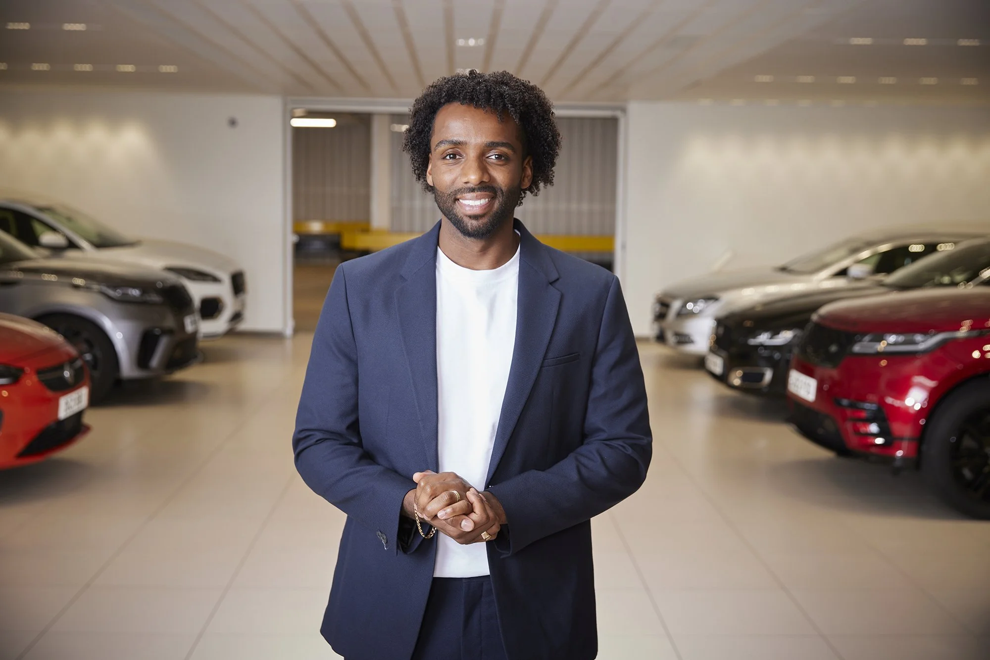 A man in a dark blue suit standing in a car showroom with several cars parked behind him.