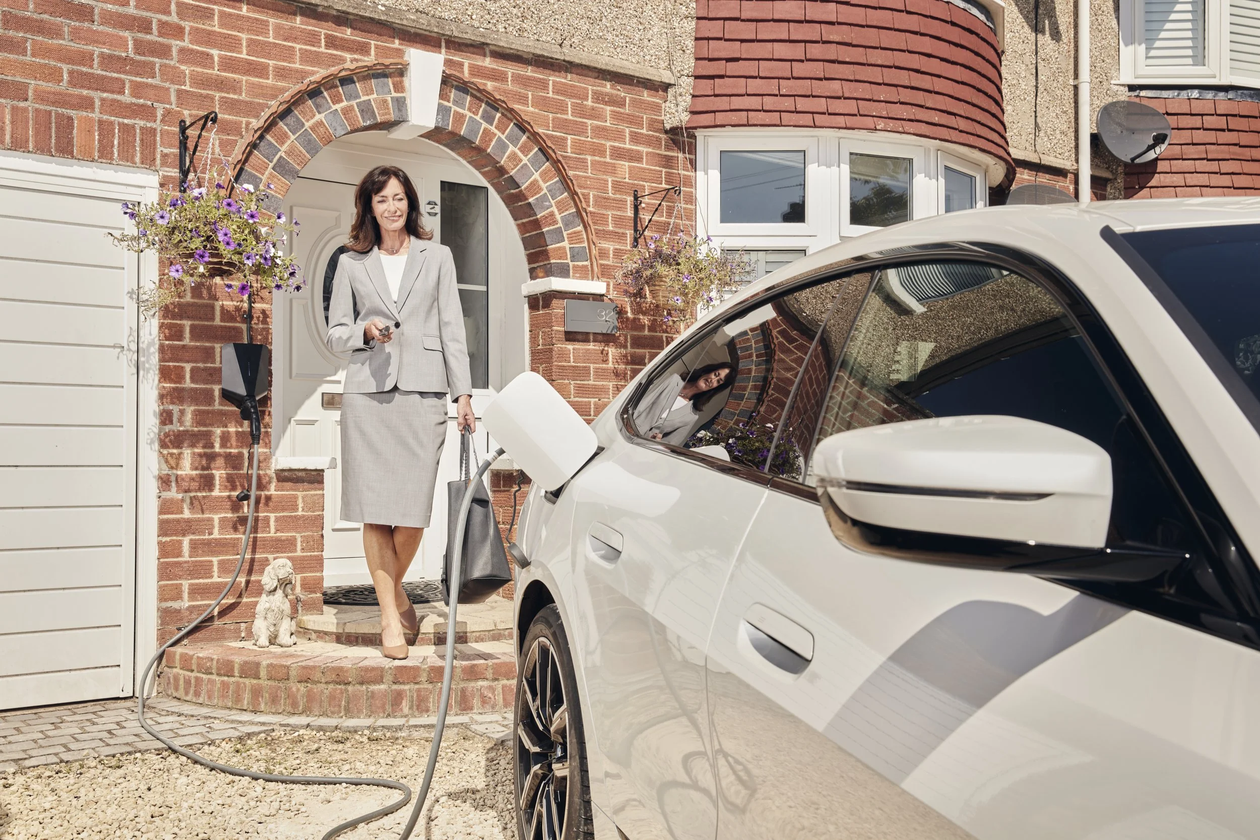 A woman in a gray business suit charging an electric car outside a brick house