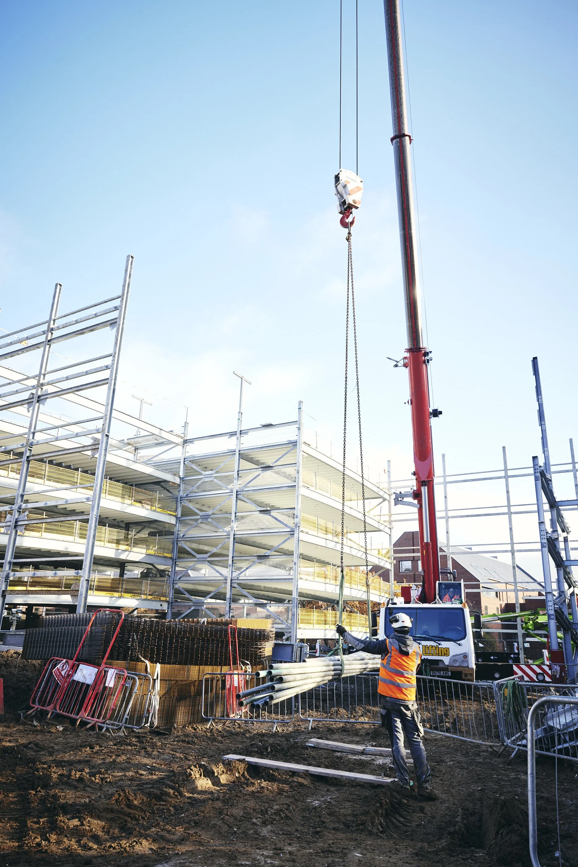 A construction site with a worker wearing a helmet and safety vest standing near a crane lifting pipes with a metal structure in the background.