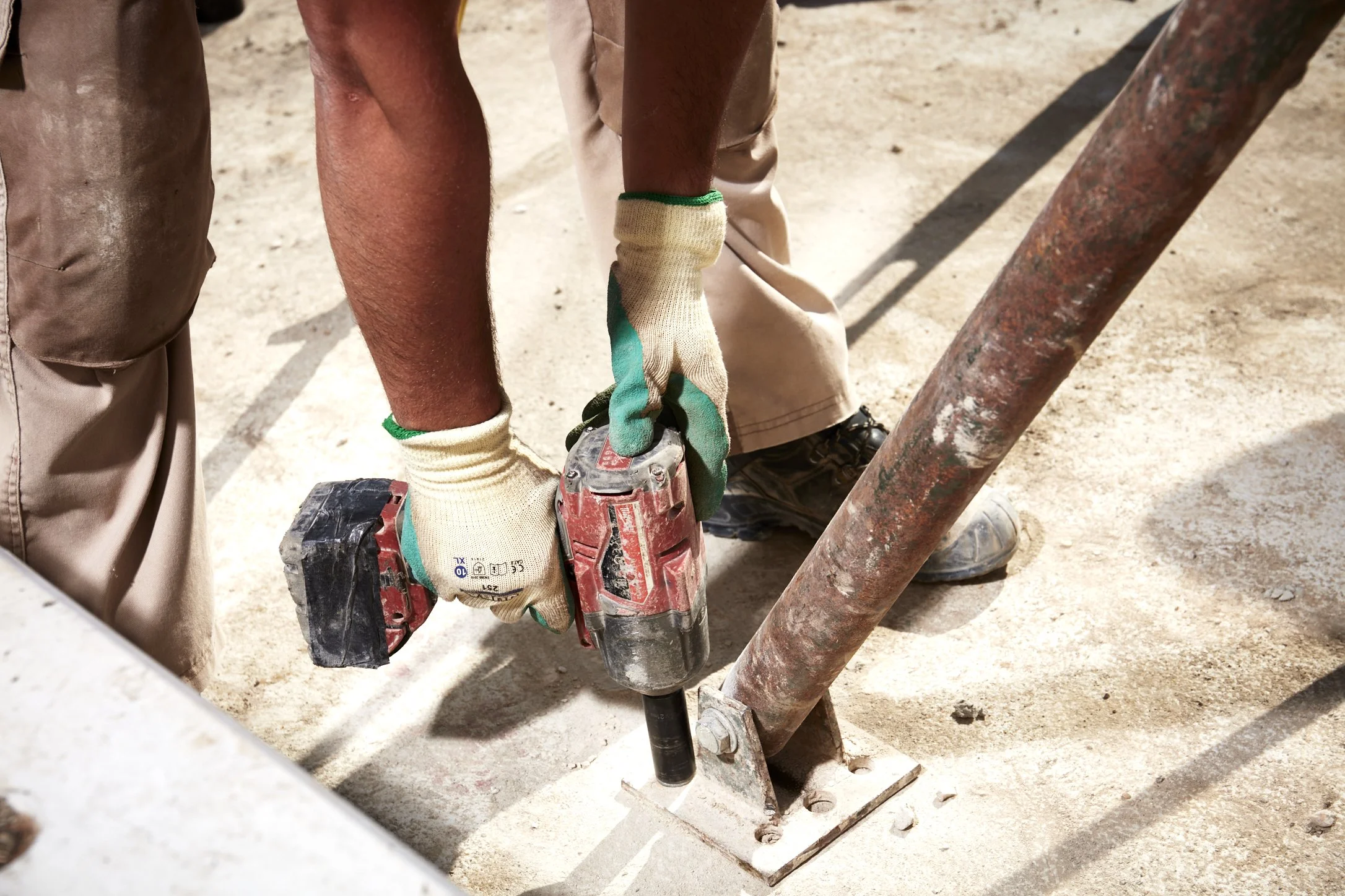 Worker using a cordless drill to anchor a metal pipe fixture to the ground on a construction site.