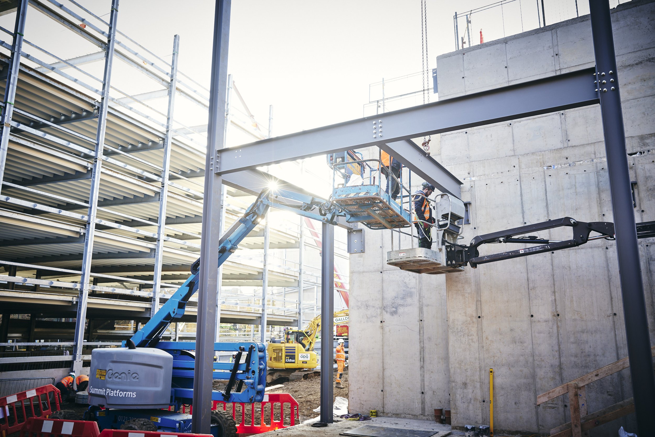 Construction workers operating a lift to work on a large concrete wall, with scaffolding and construction equipment around.