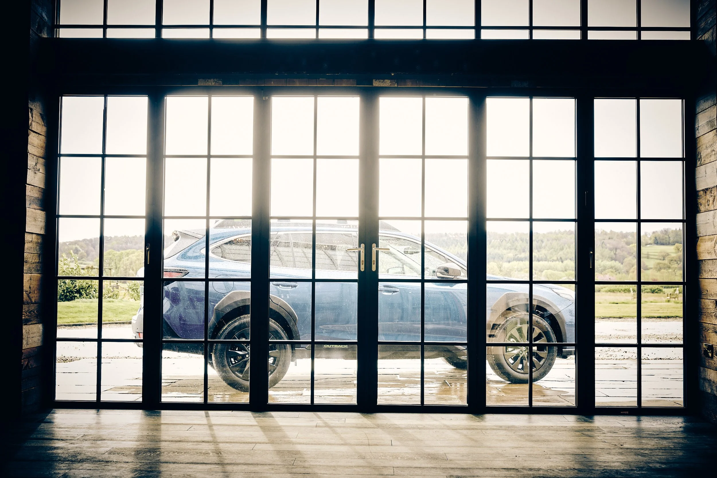 View through large glass sliding doors shows a blue SUV parked outside on a wooden porch with a rural landscape of fields and trees in the background.