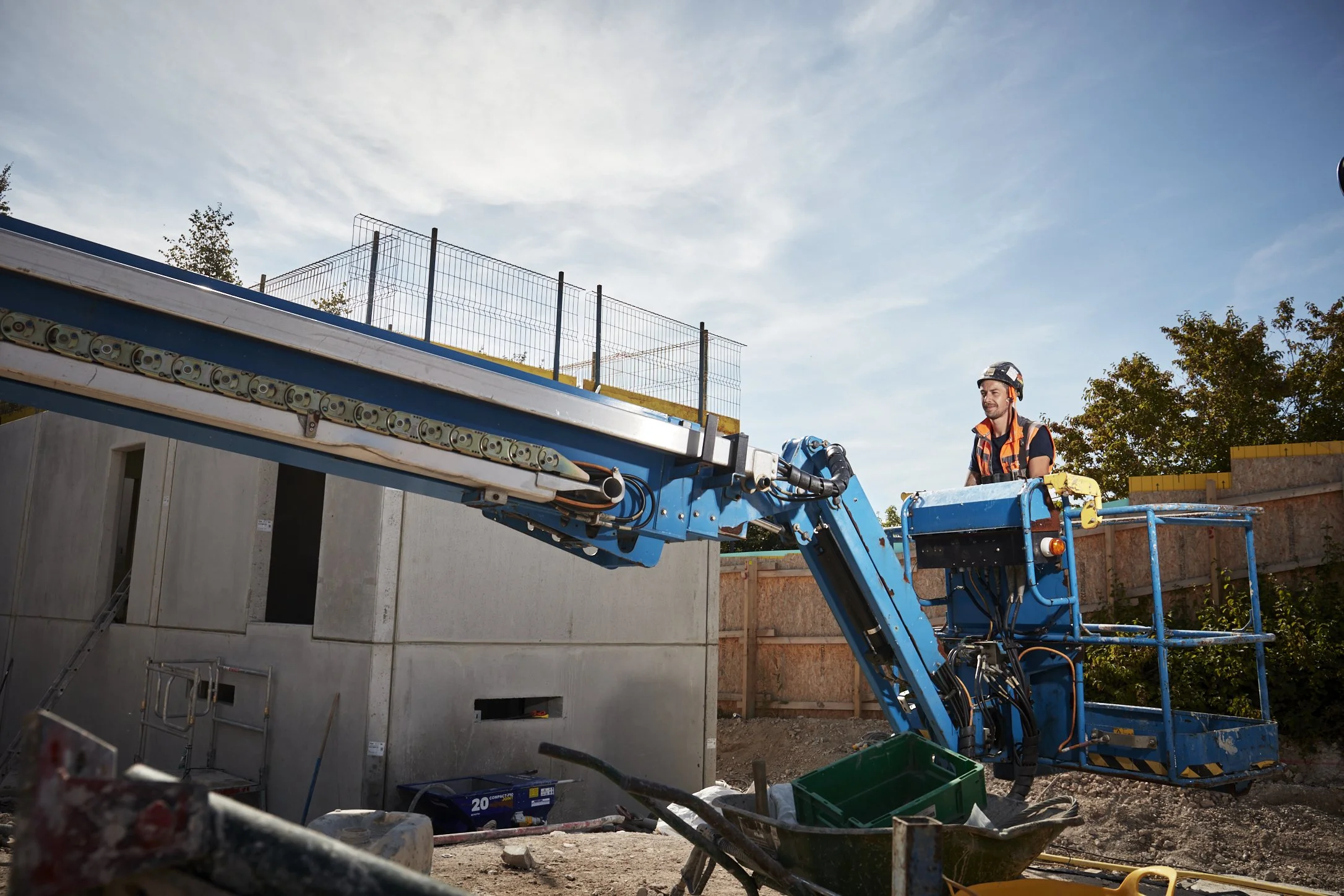 Construction worker on blue aerial lift operating near a building at a construction site with wooden fencing and trees in the background.