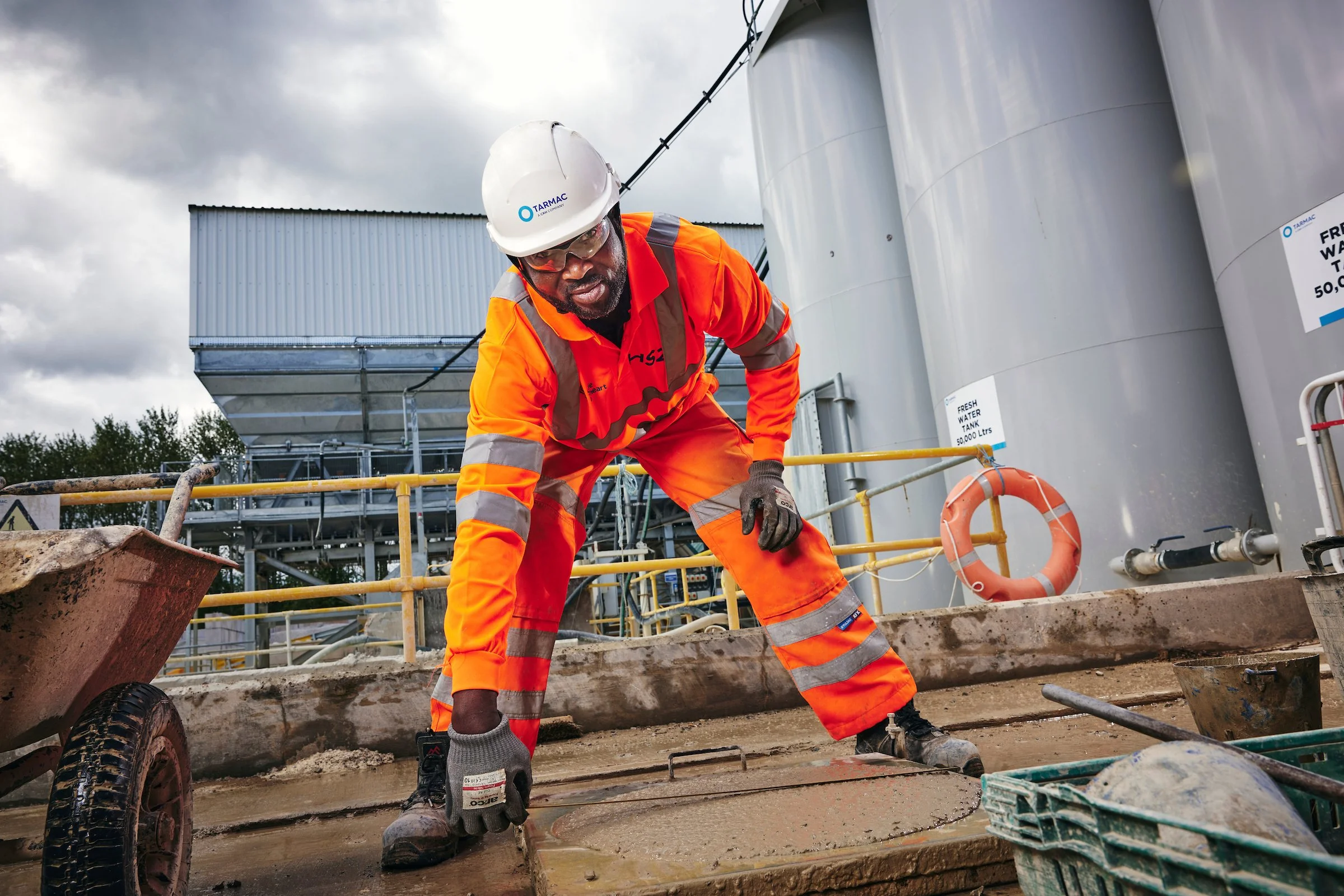 Construction worker wearing orange safety gear and helmet, bending down on a construction site with large industrial tanks and equipment in the background.