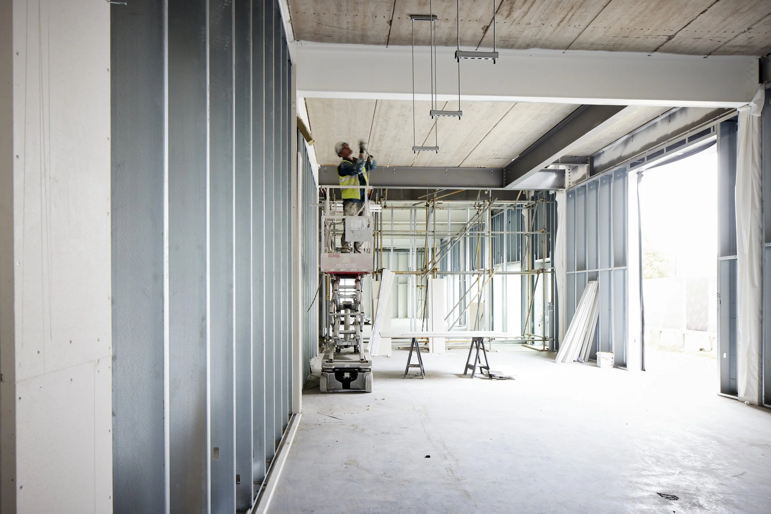 A construction worker in a yellow safety vest and helmet is working on the ceiling inside a building under construction, with scaffolding and partial wall framing around him.