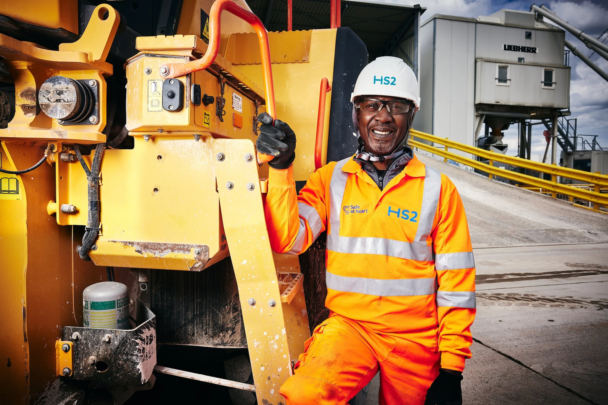 A smiling construction worker wearing orange safety gear, a white helmet with 'HS2' written on it, and safety glasses, standing next to a yellow construction machine at a construction site.