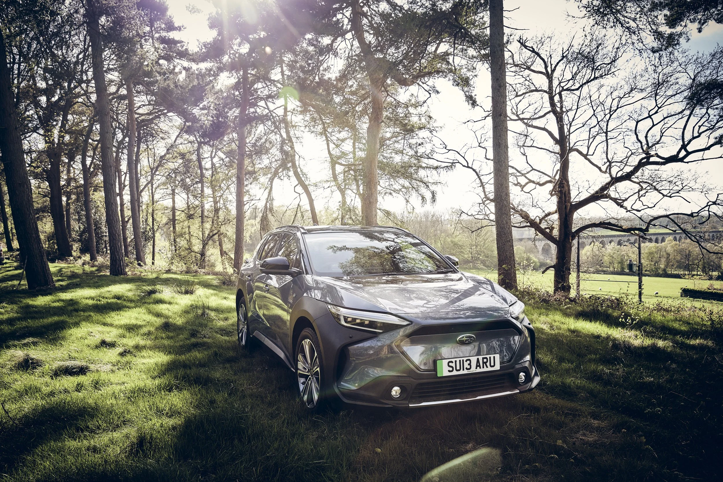 A gray hatchback car parked on grass in a wooded area with trees and sunlight shining through.