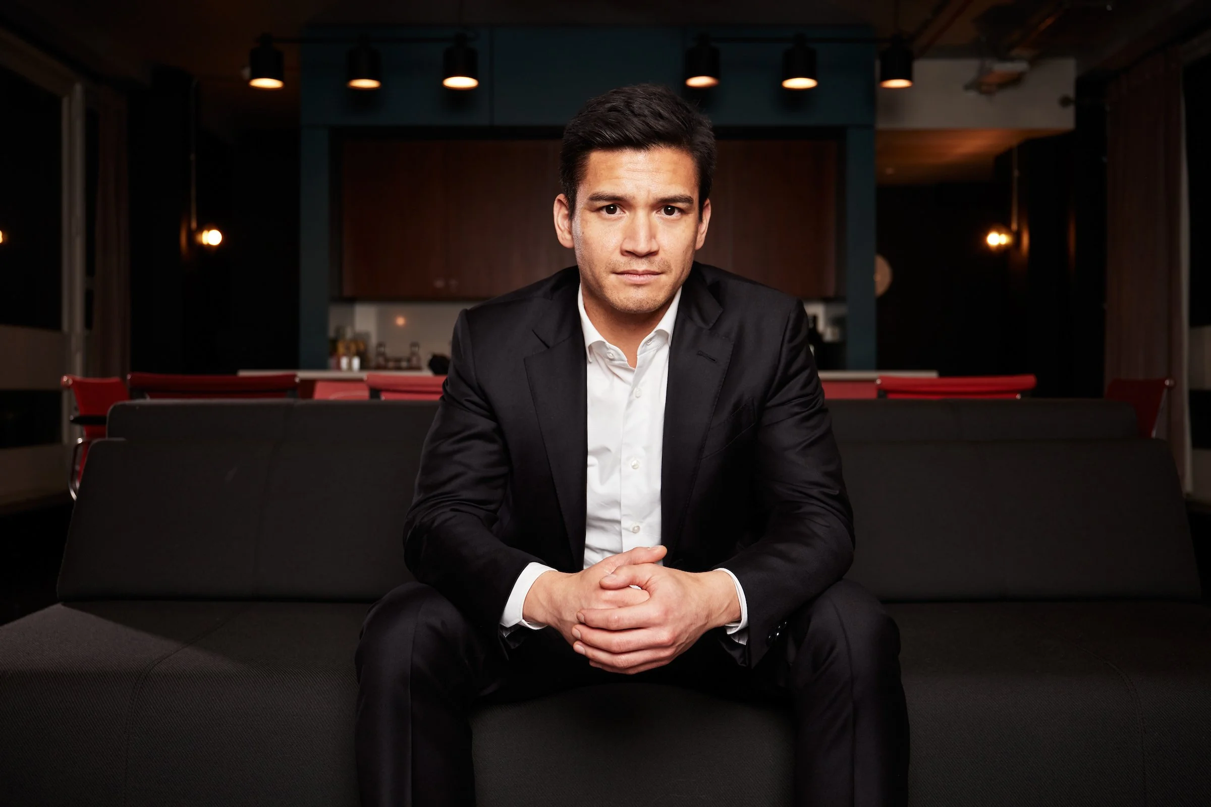 A man in a black suit and white shirt sitting on a black couch in a modern, dimly lit restaurant or lounge.