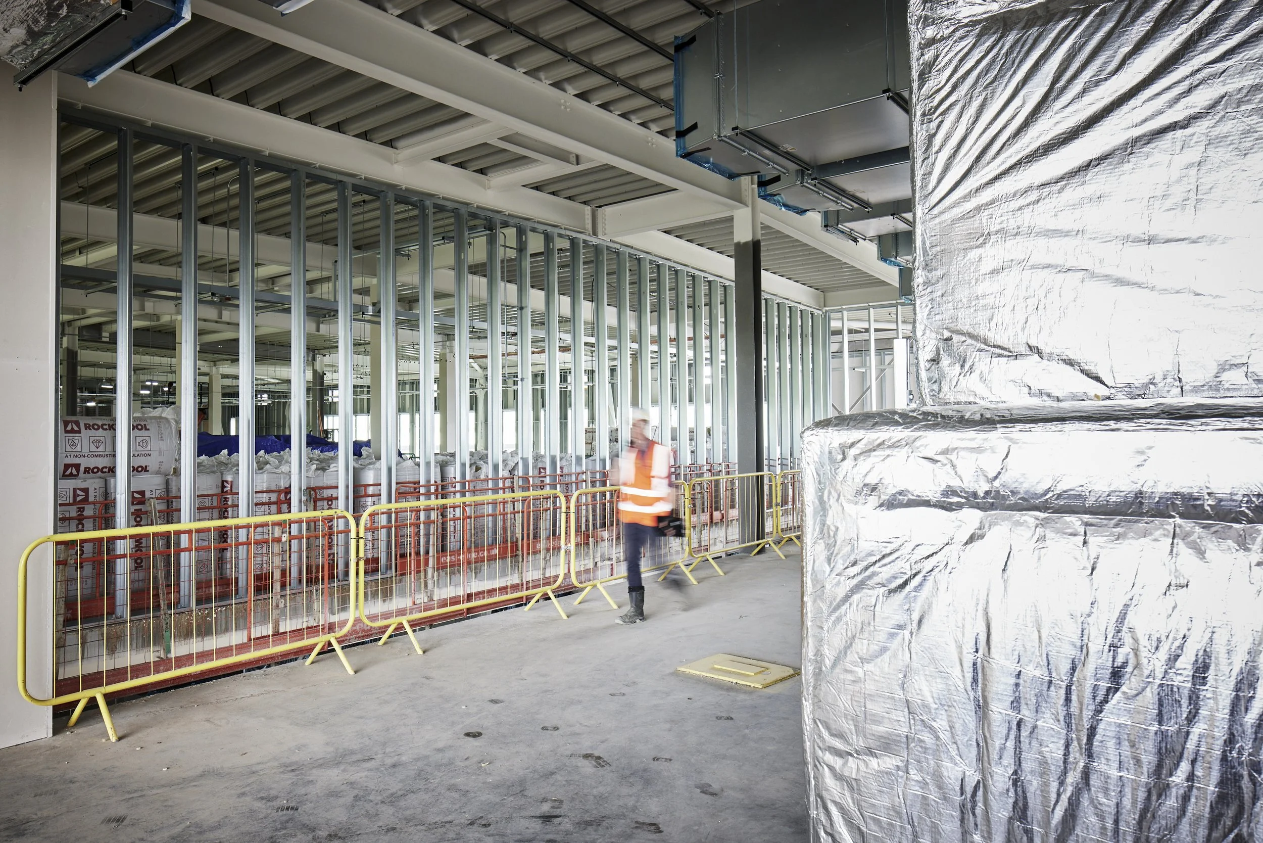 Construction worker in an orange safety vest and black work boots standing inside a building under construction, with metal framing for walls, insulation, and ductwork visible, and yellow safety barriers along the floor.