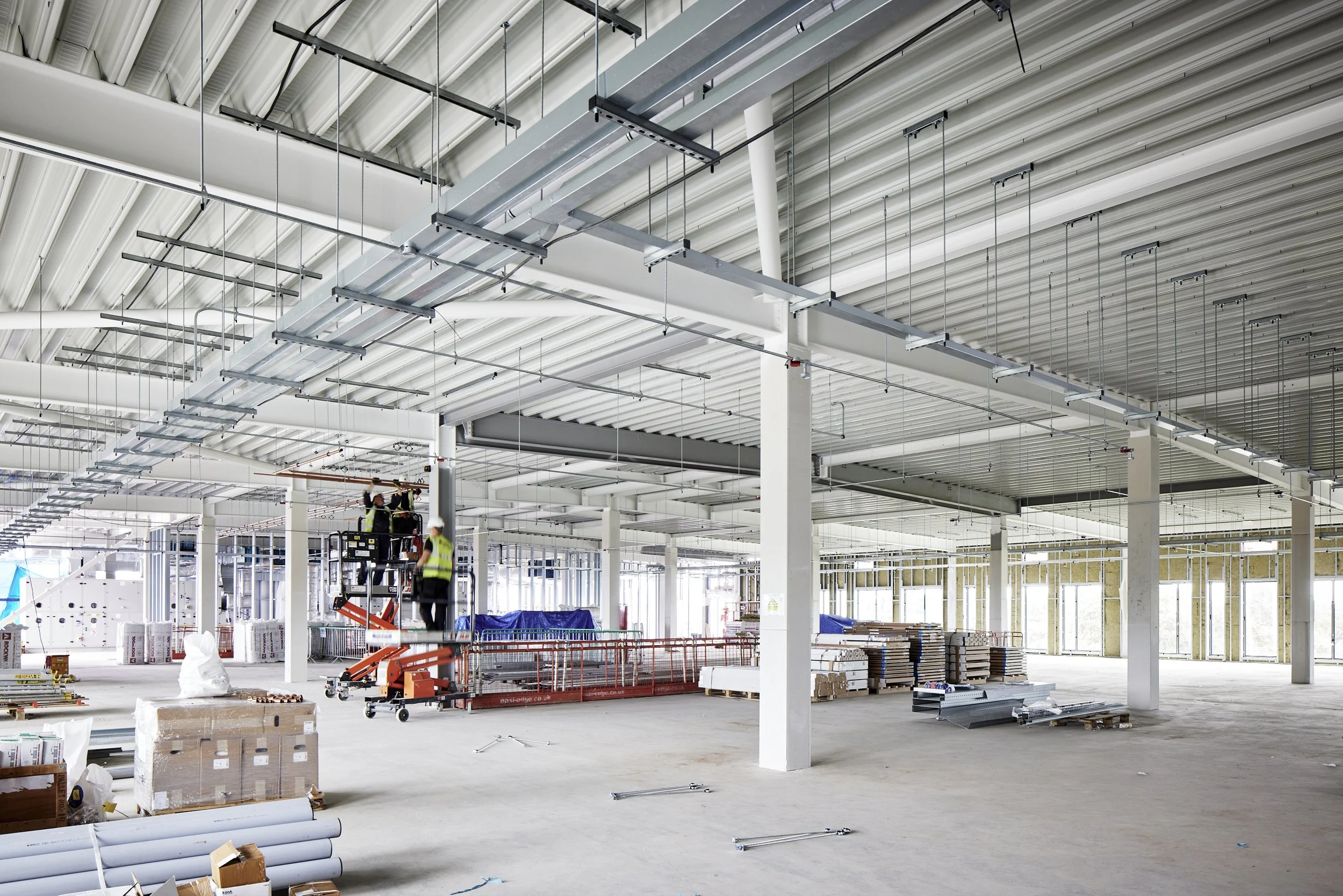 Construction workers installing ceiling ductwork inside a large, open commercial or industrial building with unfinished walls and floor, construction materials, and equipment scattered around.