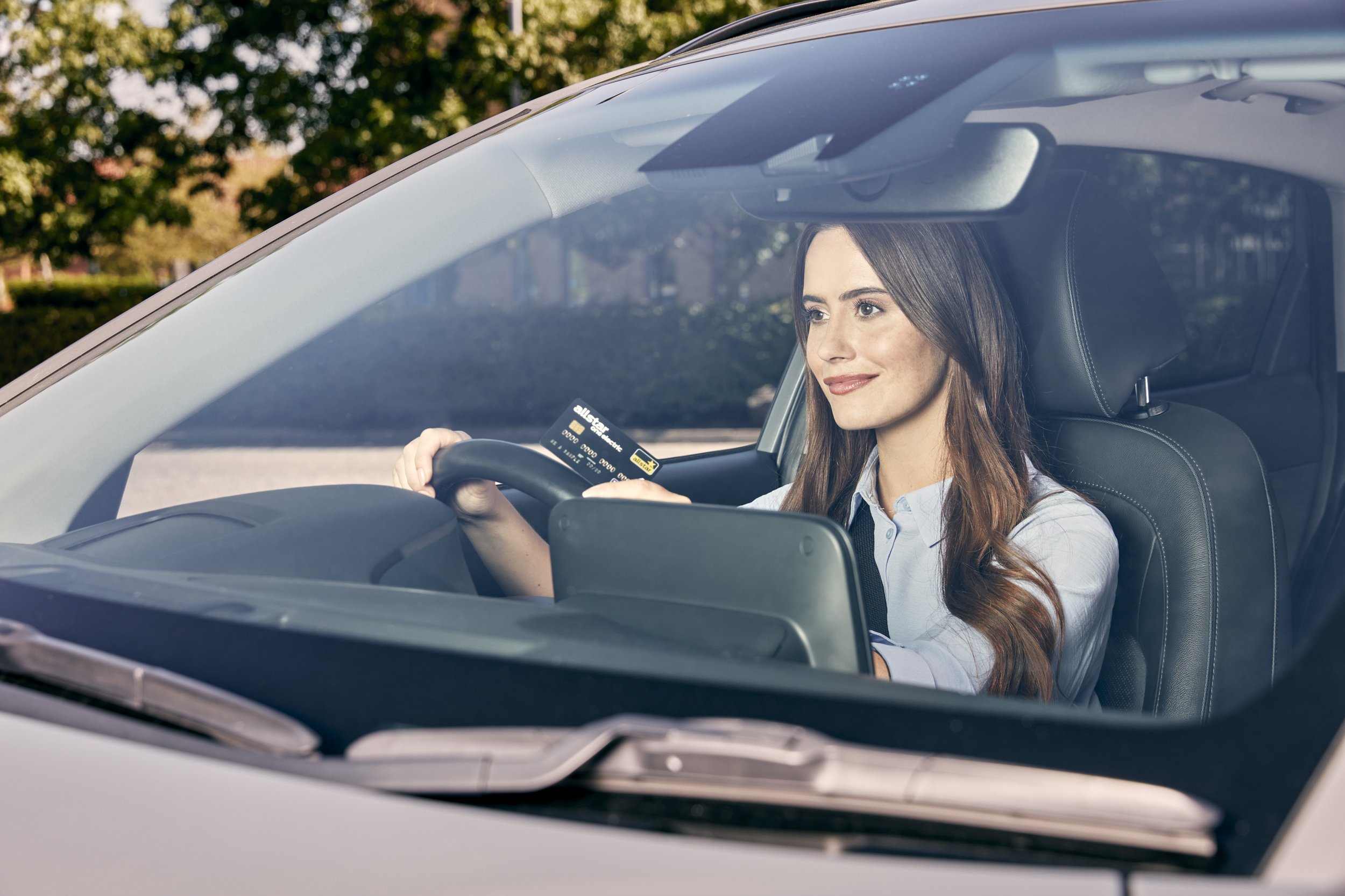 A woman with long brown hair driving a car, holding a credit card in her right hand and a tablet in her left, looking out the windshield with a smile.