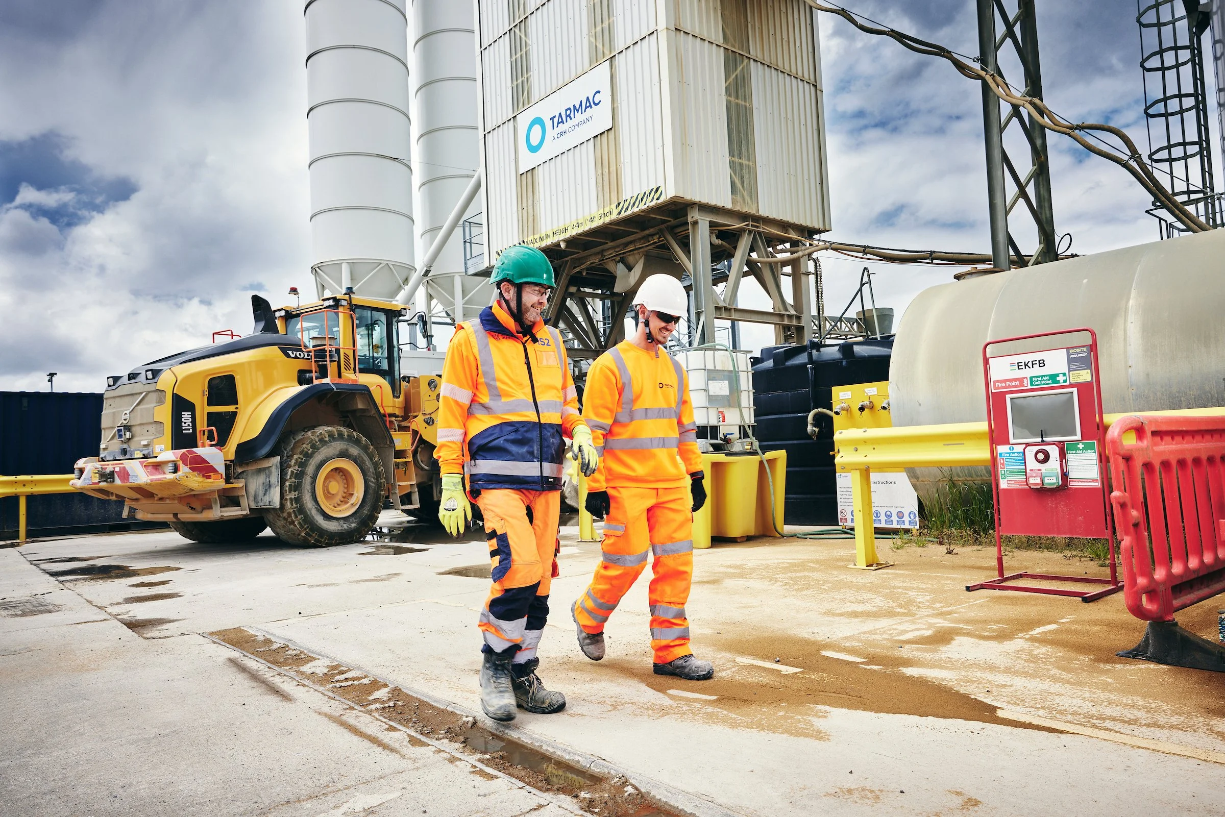 Two construction workers in orange safety gear walking on a construction site with equipment and machinery, including a large yellow vehicle, storage tanks, and safety signs in the background.