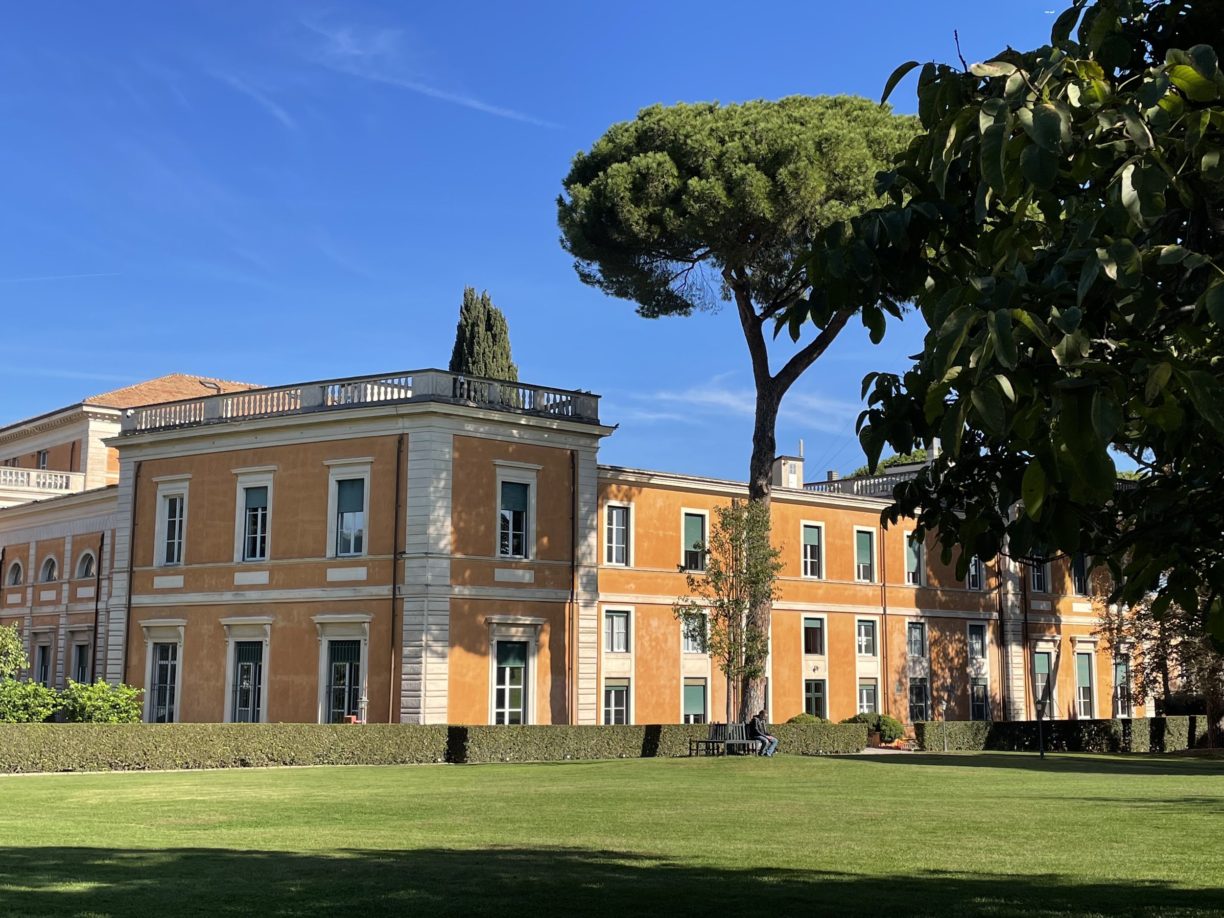 A large, orange-colored historic building with tall windows and white decorative accents, surrounded by a well-maintained green lawn, tall trees, and clear blue sky.