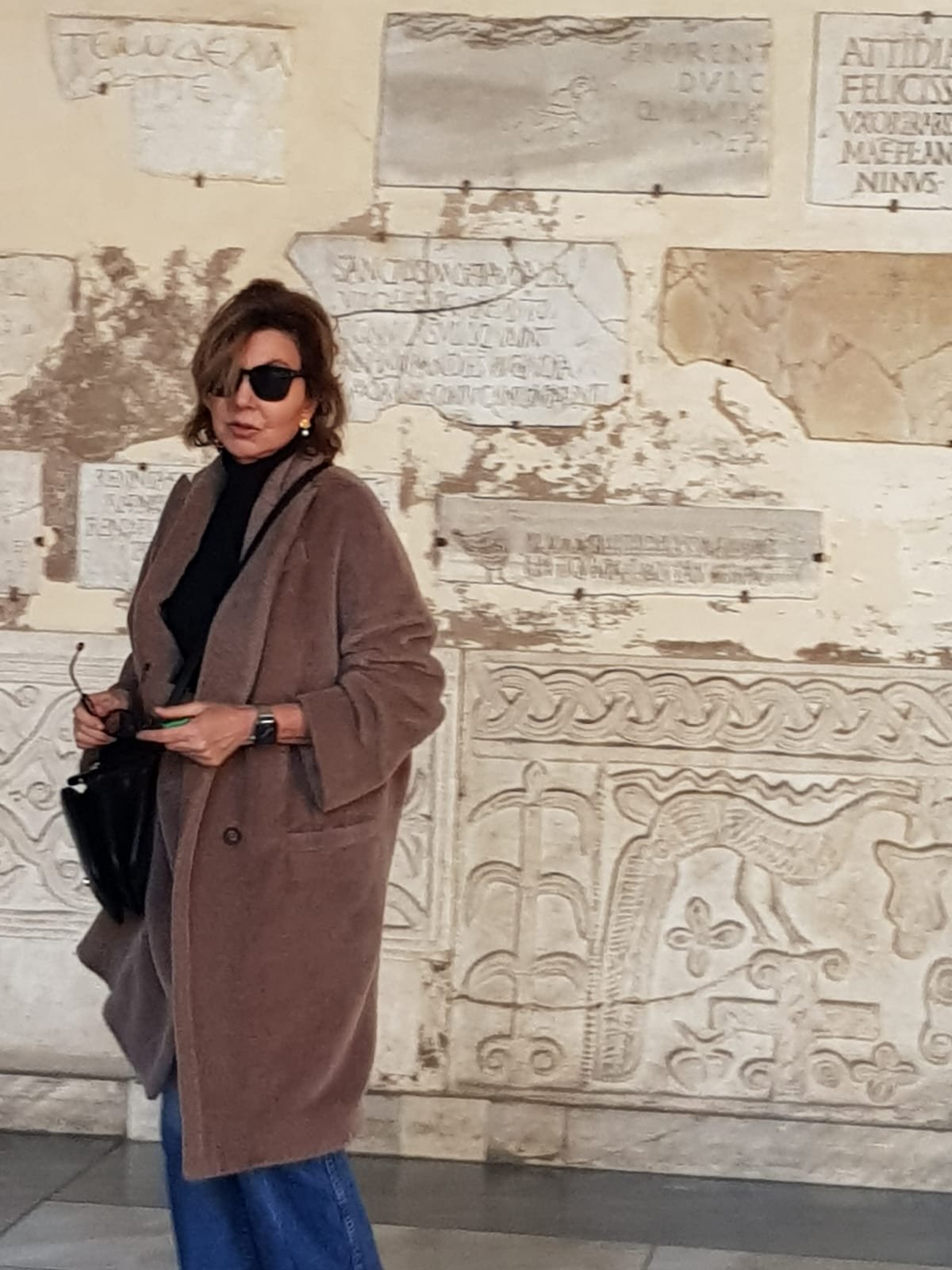 A woman in a brown coat, sunglasses, black turtleneck, and blue jeans standing in front of a wall with ancient engraved stones.