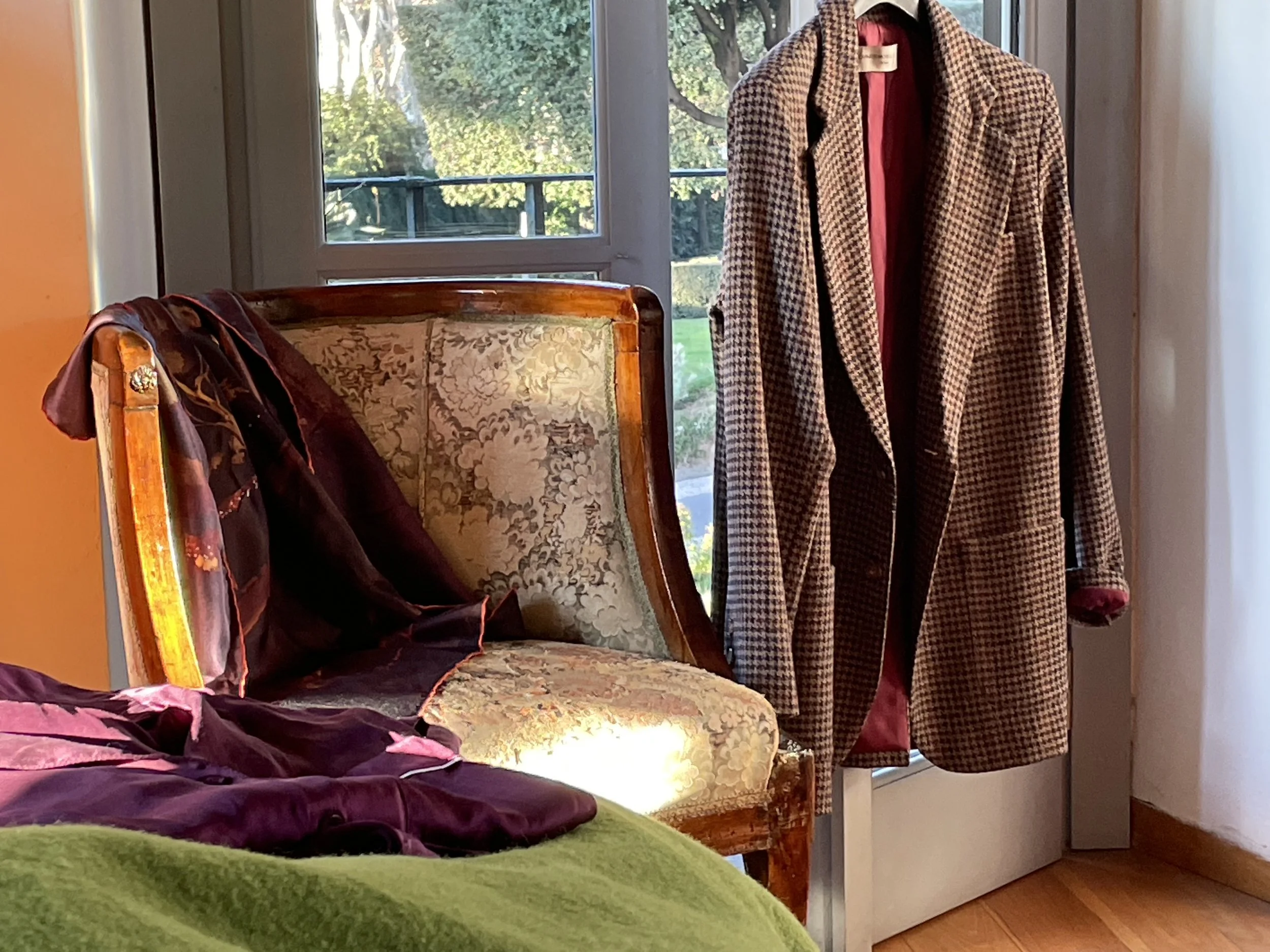 A vintage armchair with floral upholstery and a purple garment draped over it, near a window with a view of trees, with a checkered jacket hanging on a hook behind it.