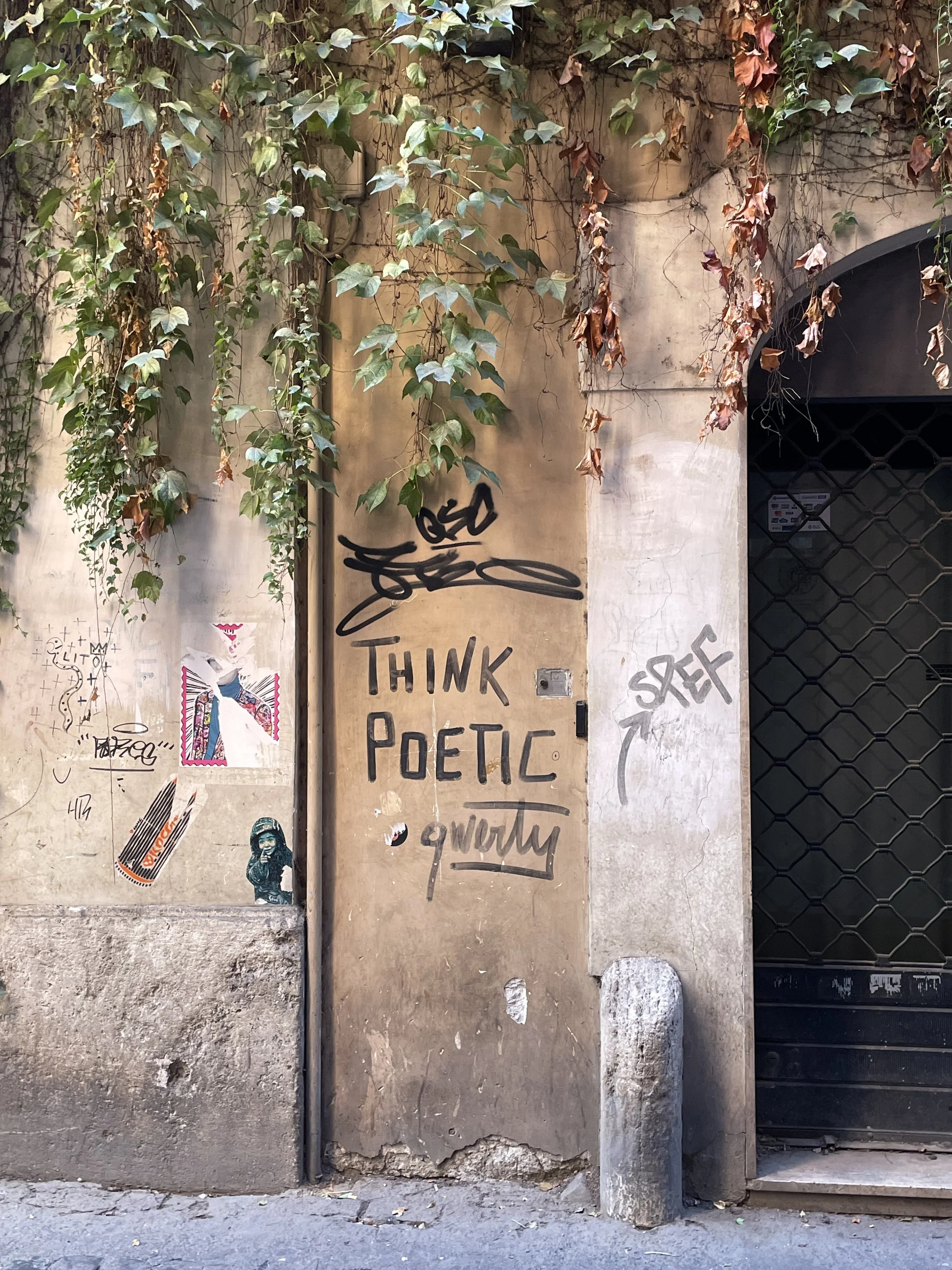 Graffiti on a beige wall reads 'Think Poetic' with smaller unreadable words below, surrounded by vines and posters, next to a black gate with a stone barrier in front.