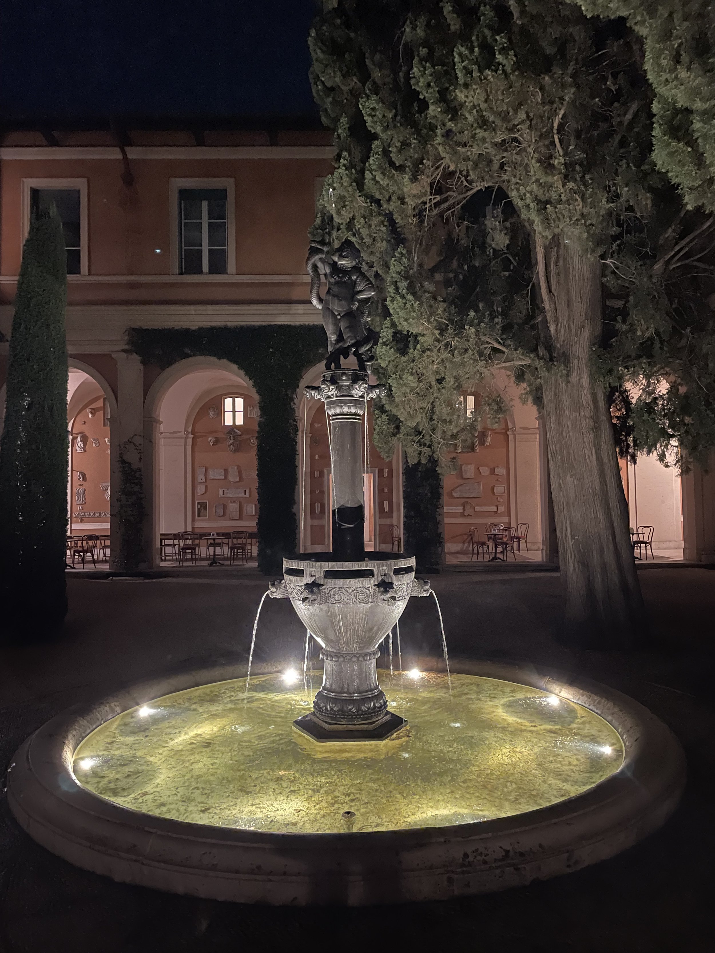 Night view of a decorative fountain with a statue on top, surrounded by trees and a building with arches and windows in the background.