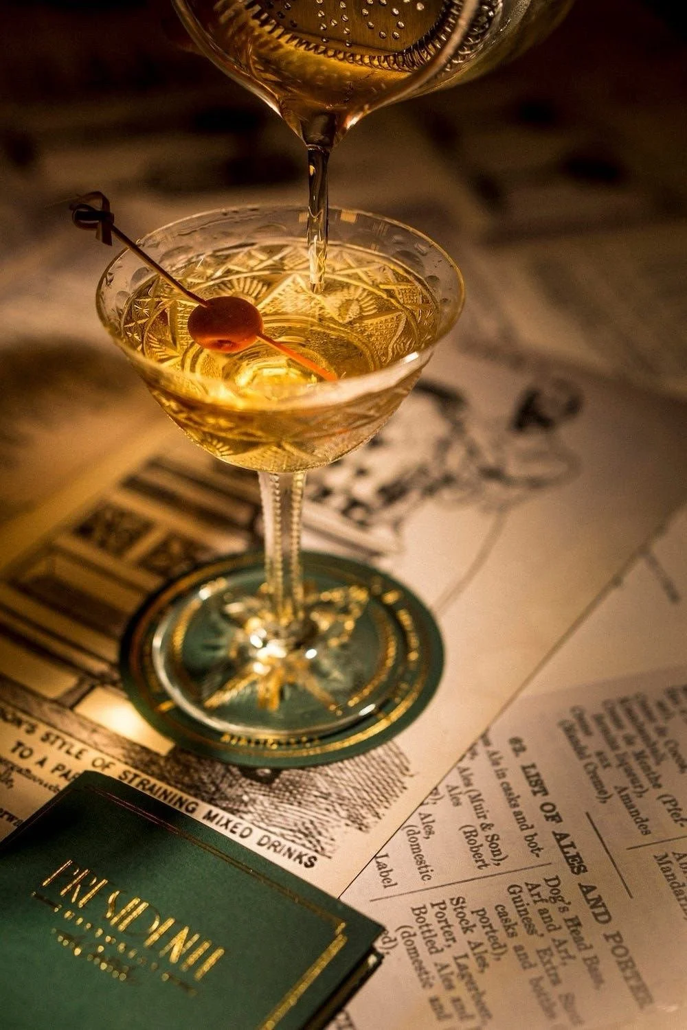 A cocktail glass with a cherry on a skewer and a drink being poured into it, placed on a table with menus and a green box with gold lettering.