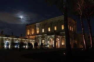 Nighttime view of a historic building illuminated with exterior lights, with palm trees in the foreground and a moon in the partially cloudy sky.