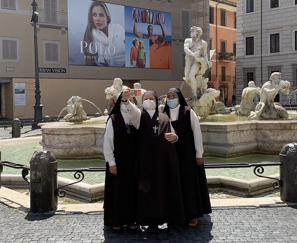 Three nuns taking a selfie in front of a fountain with sculptures, in an urban area with billboard advertisements.