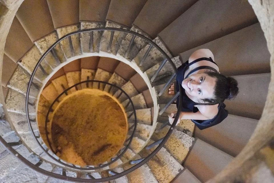 A woman with dark hair tied in a bun, looking up and smiling, is standing at the top of a spiral staircase viewed from above.
