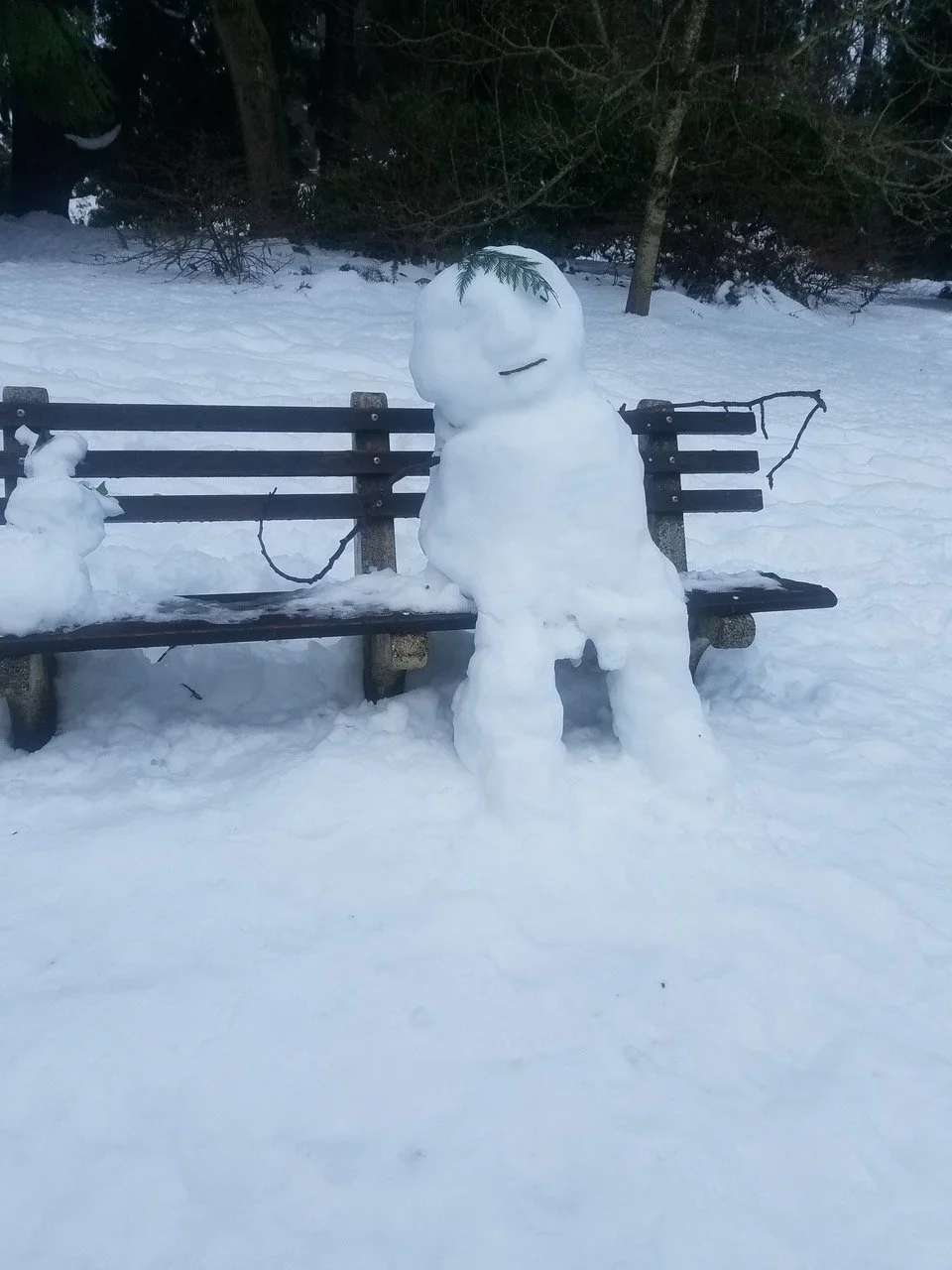 A snow sculpture resembling a snowman with a large rounded head and body, with a branch sticking out as an arm, sitting on a park bench in a snowy landscape with trees in the background, and a small snow sculpture on the left.