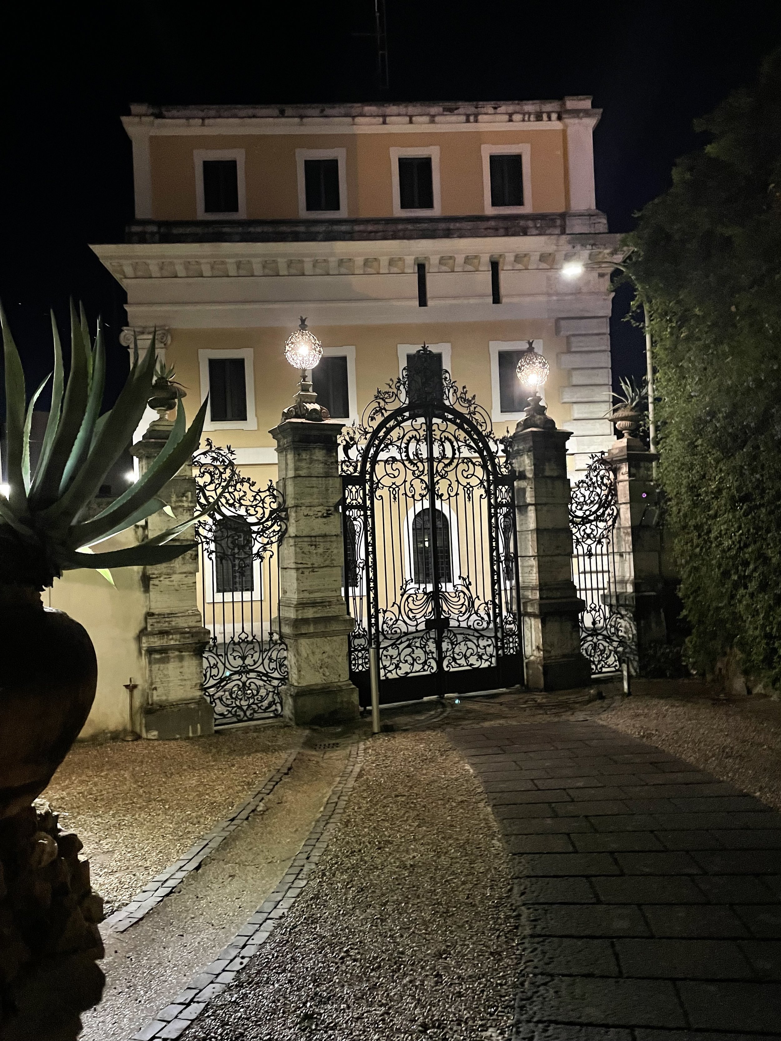 A large, ornate metal gate in front of a grand, three-story mansion at night. The mansion has a yellow facade with white accents, multiple windows, and decorative details. The gate is flanked by stone pillars and topped with spherical lights. There i