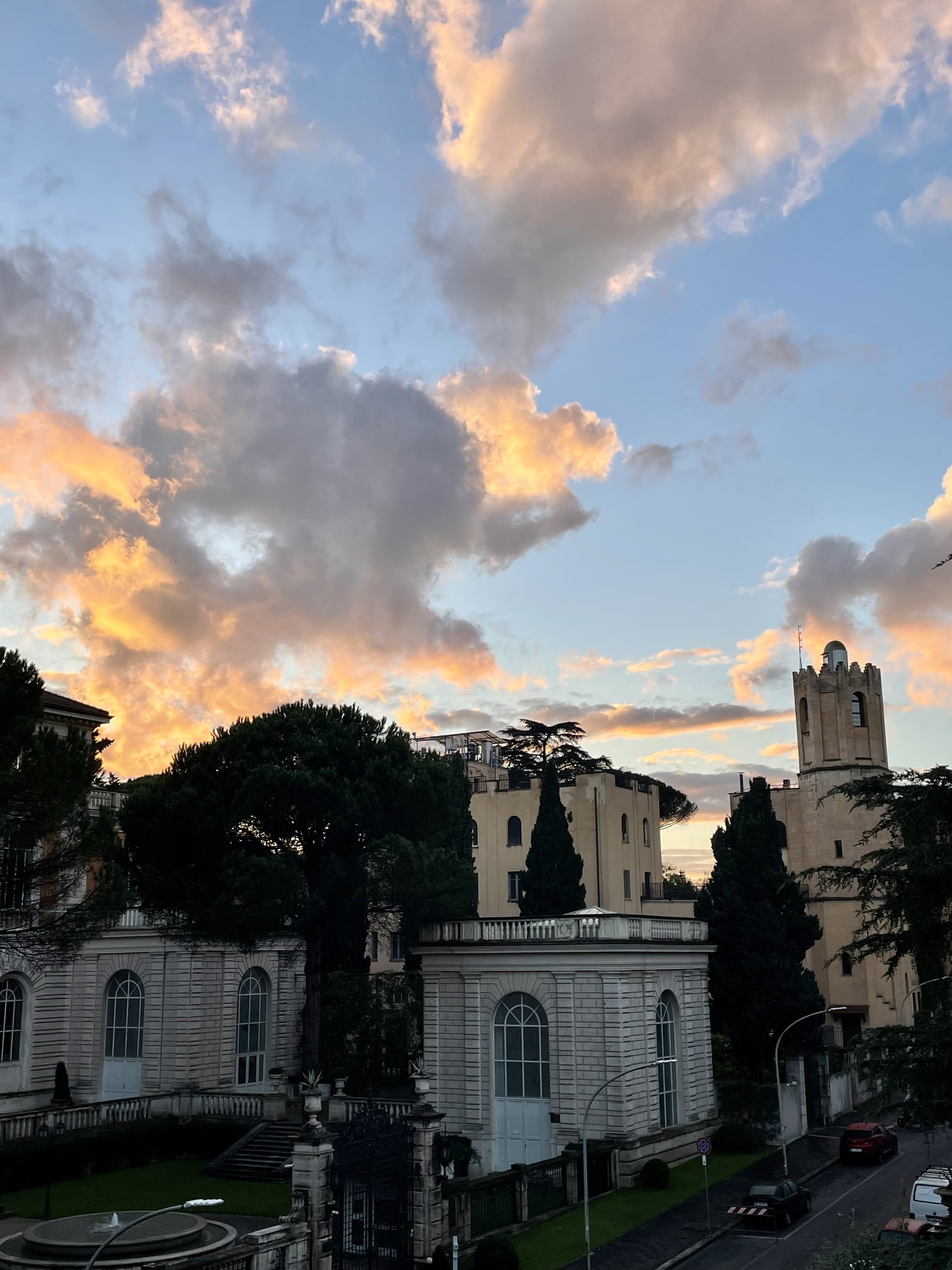 Sunset sky with orange and pink clouds over historic buildings and trees in a city street.