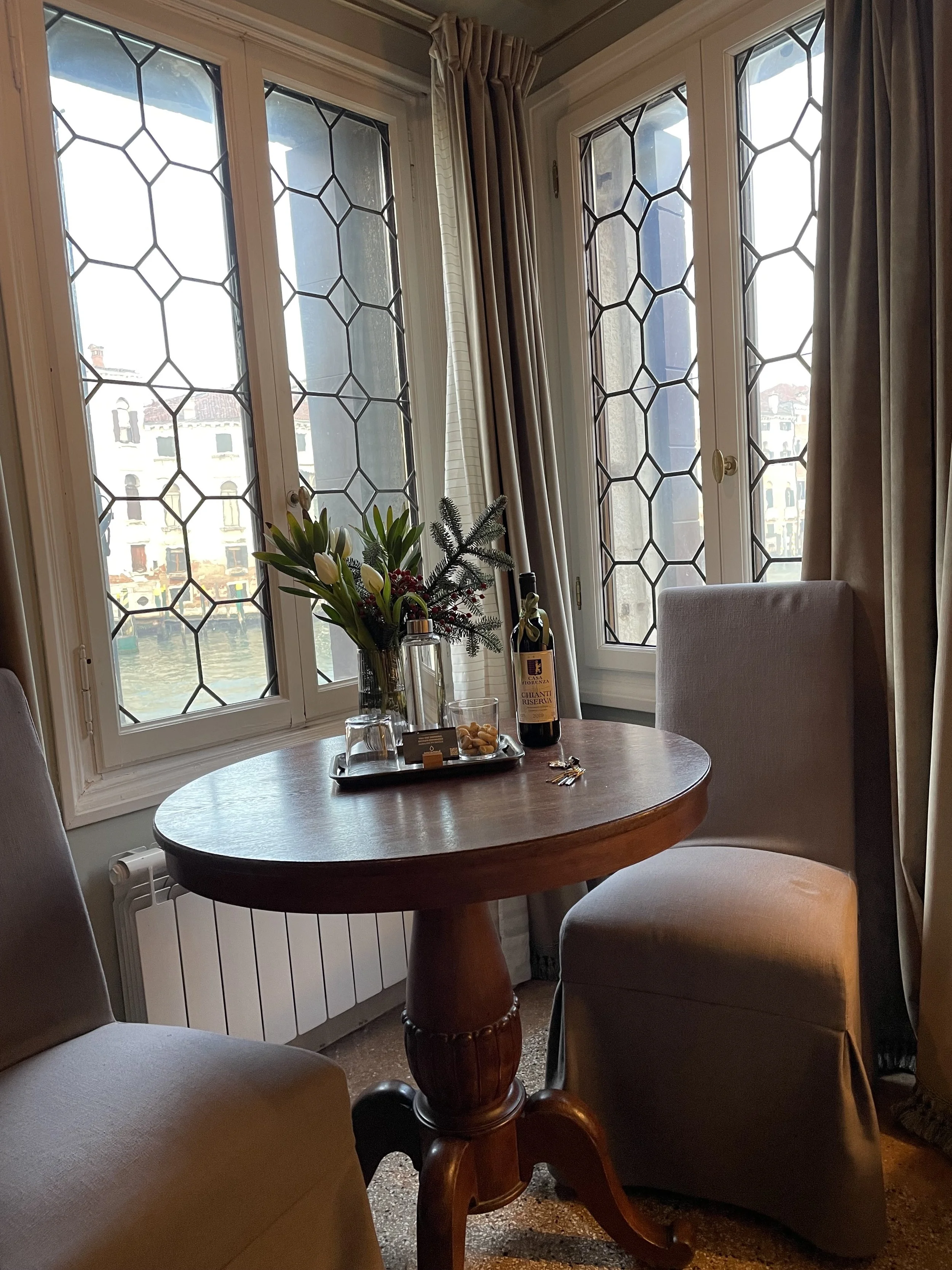 A cozy corner in a restaurant or cafe with a wooden round table, two cushioned chairs, and large windows with decorative hexagonal iron grilles, revealing a canal and buildings outside. The table has a flower arrangement, a bottle of red wine, and so