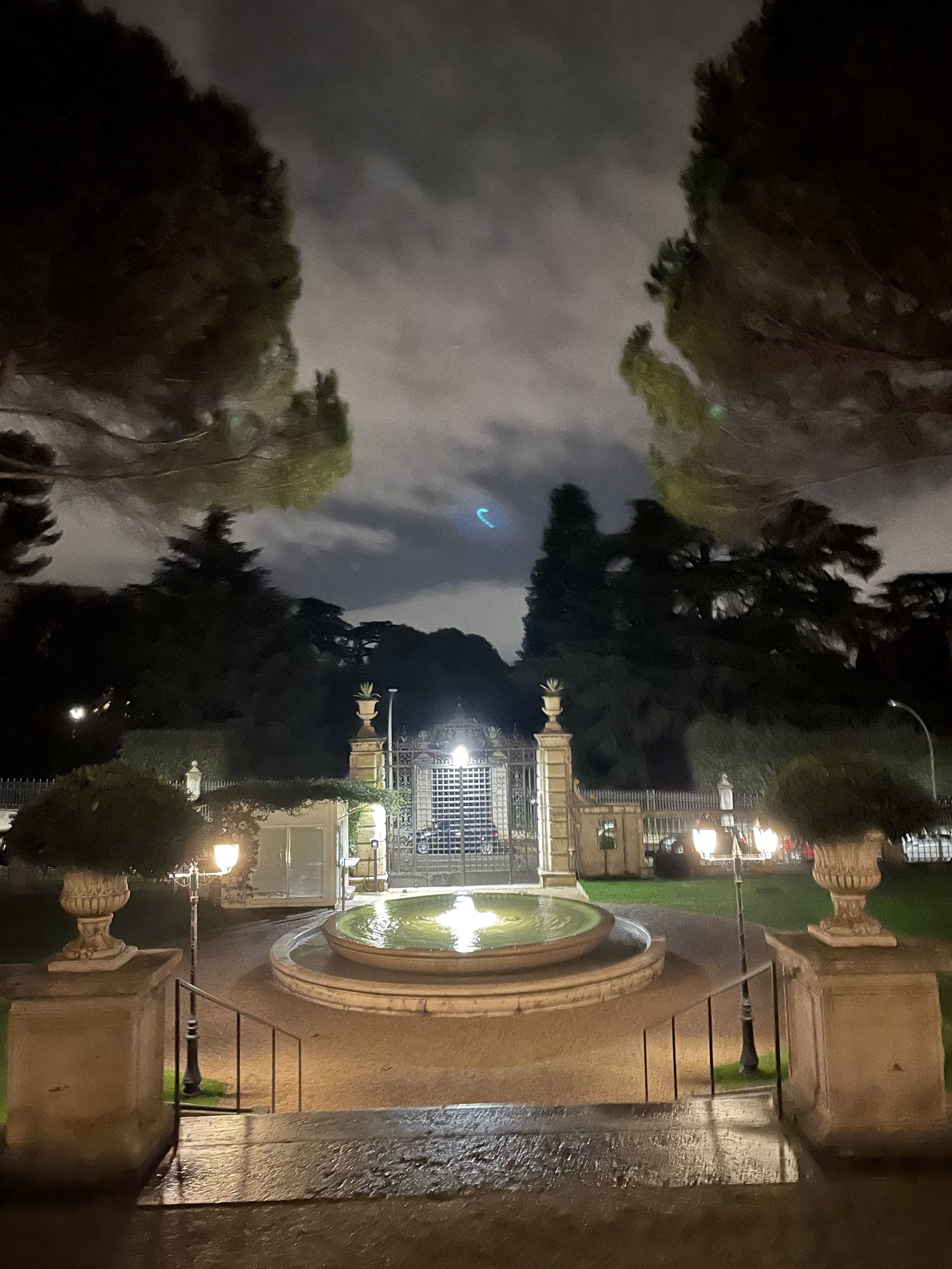 Night view of a gated estate with a fountain in the foreground, surrounded by potted plants and lampposts, under a cloudy sky.