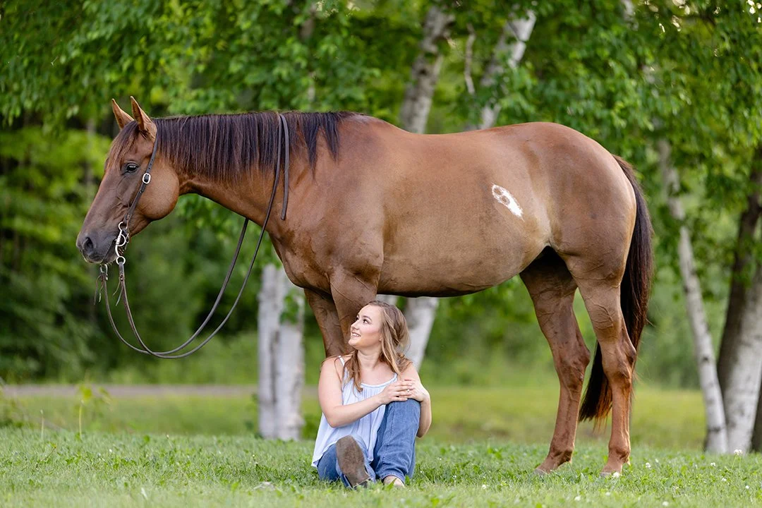 Young woman sitting in the grass beside her chestnut horse during a peaceful horse and rider portrait session surrounded by trees in Wisconsin.