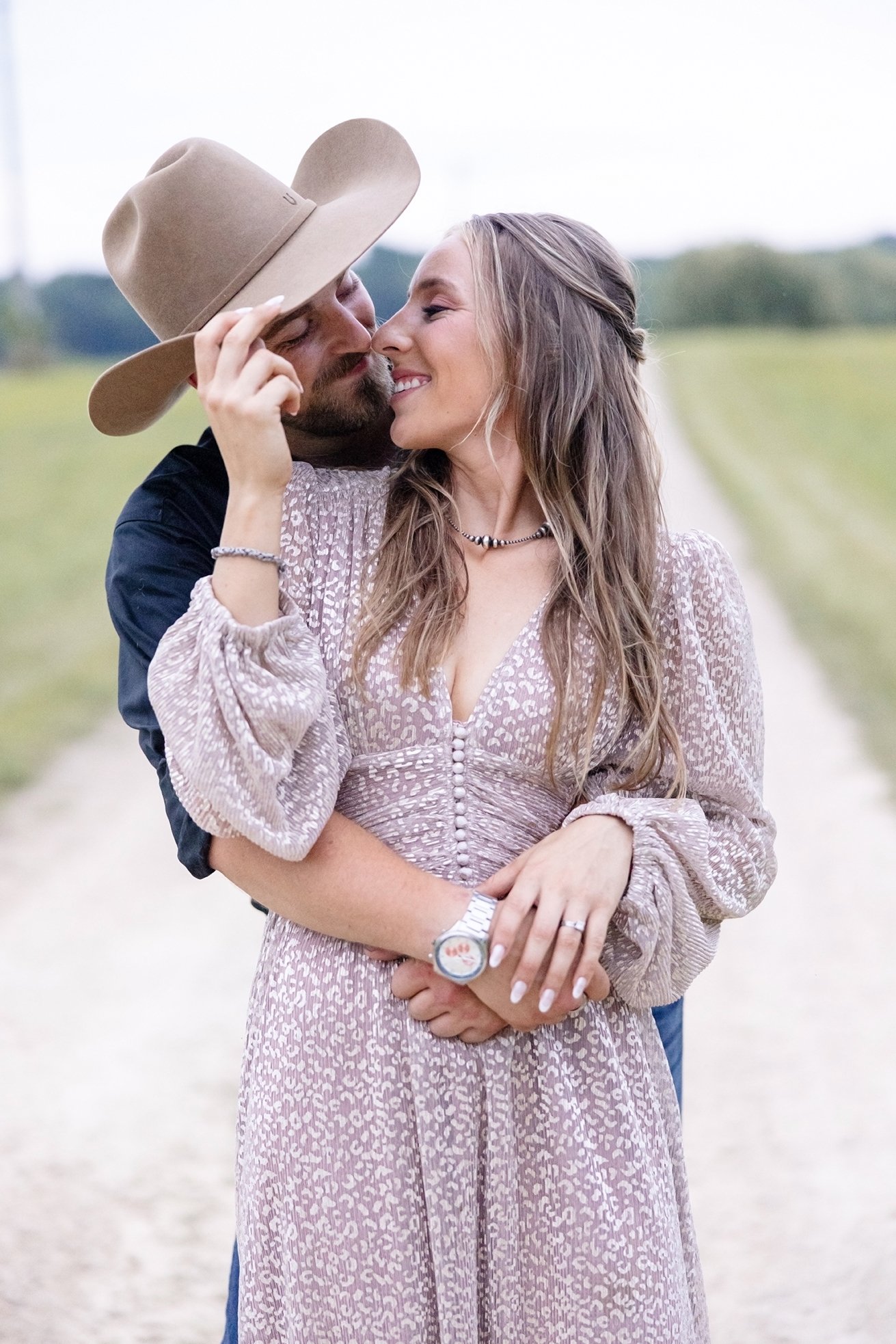 Amber tips Isaacs cowboy hat while embracing Amber during their western engagement photos on their farm in Poynette, Wisconsin, capturing a tender and playful moment.