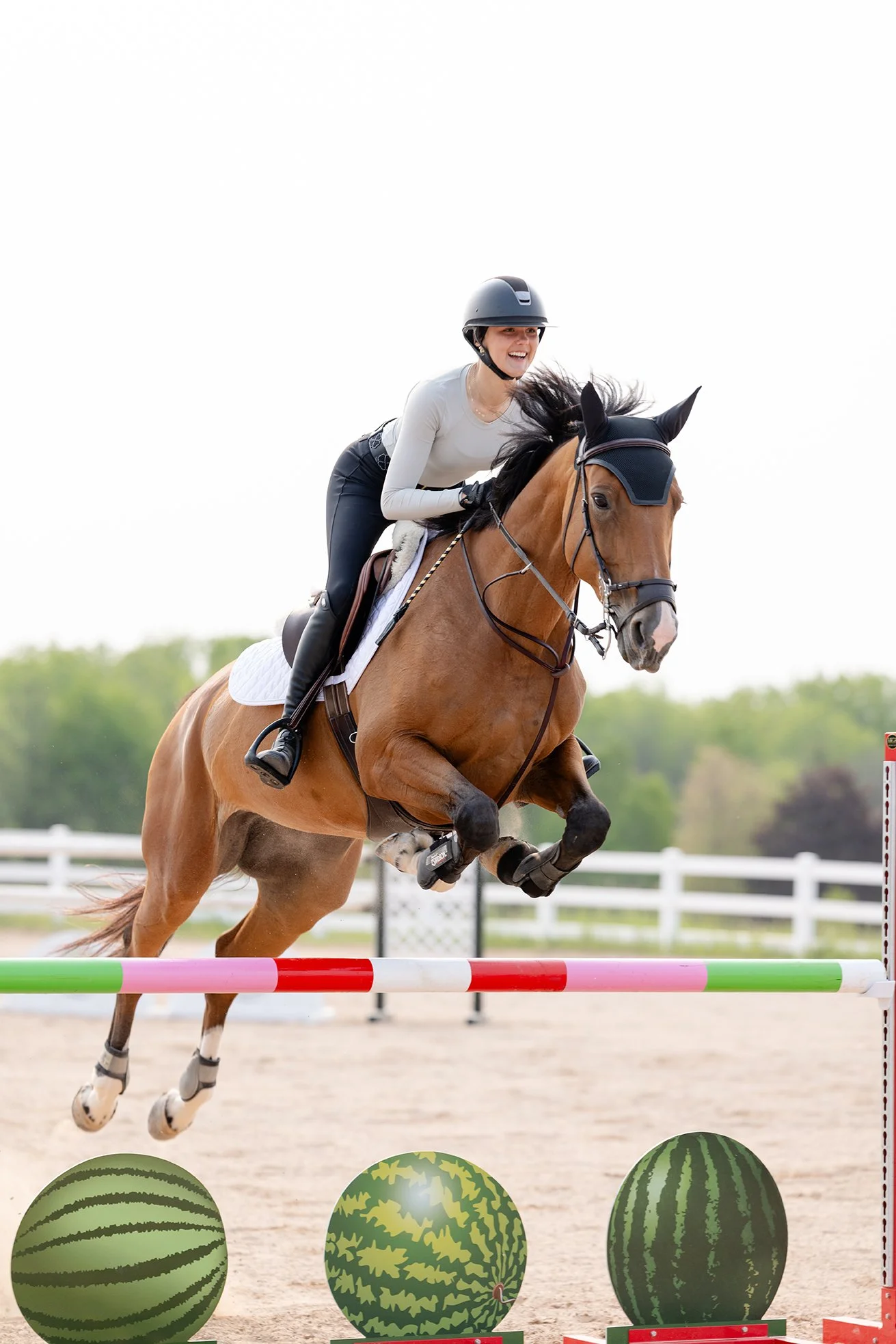 Rider in grey STS Equestrian shirt smiling while jumping a bay horse over a watermelon-themed jump.