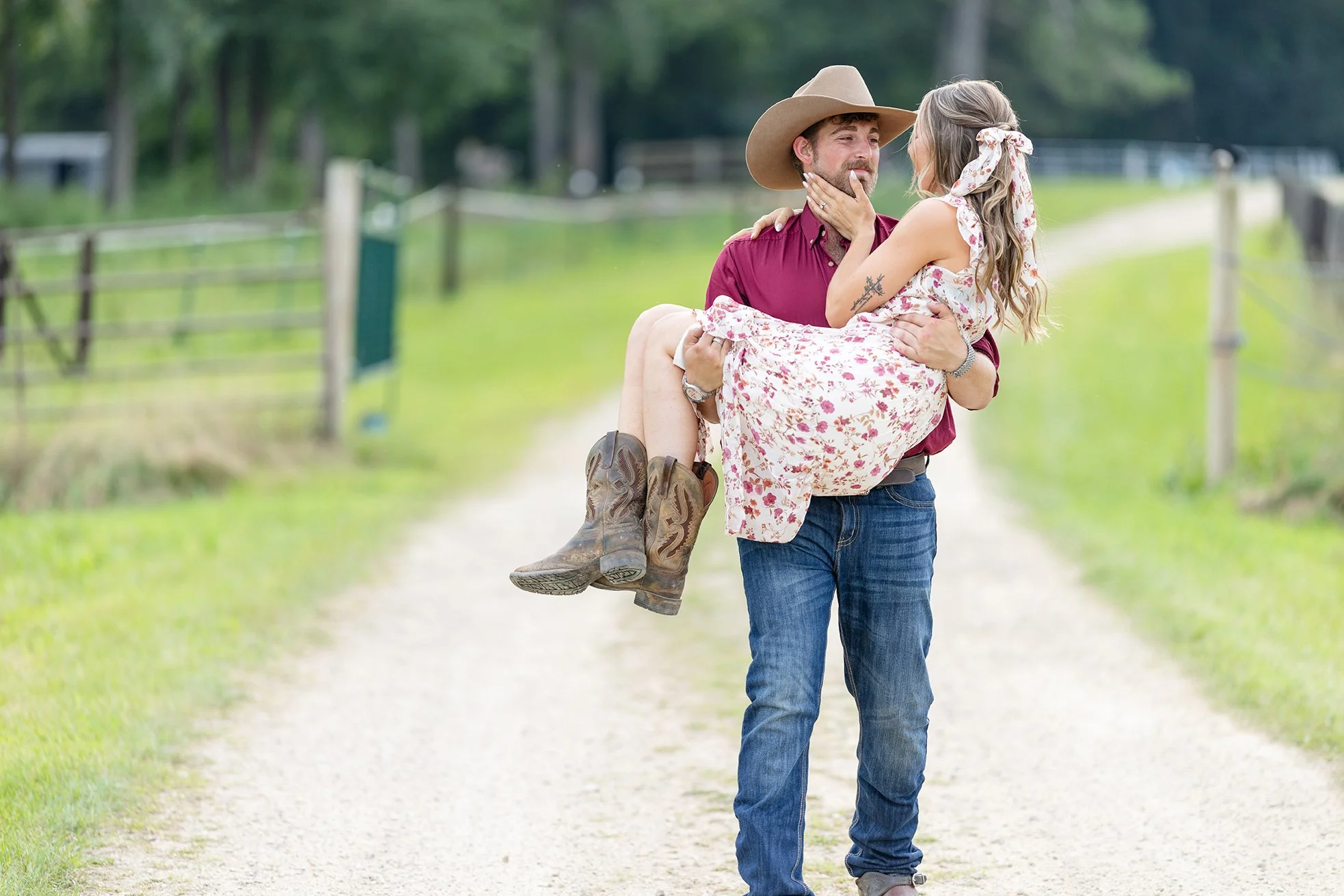 Isaac carries Amber down a gravel driveway on their Poynette, Wisconsin farm, creating a romantic moment during their western equestrian engagement portraits surrounded by green pastures and fencing.