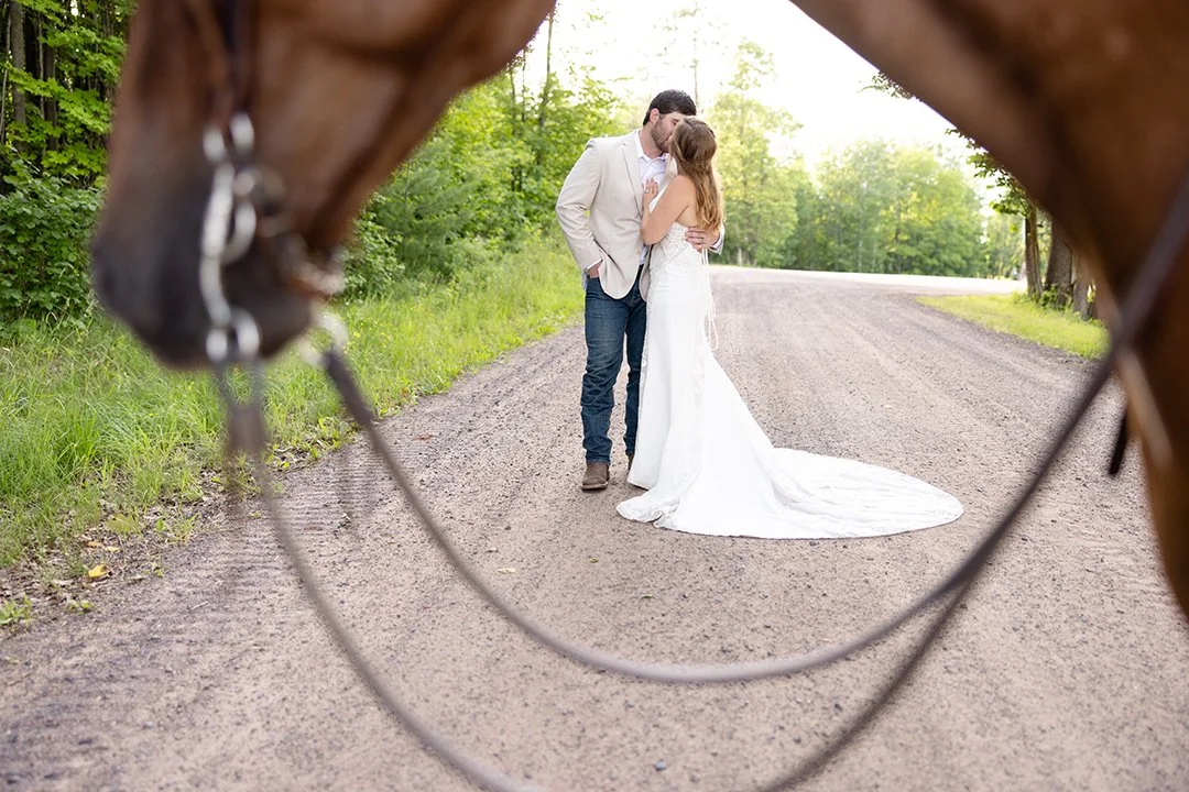 Couple kissing during their equestrian bridal portrait session framed creatively between their horse’s bridle on a quiet country road in Wisconsin.