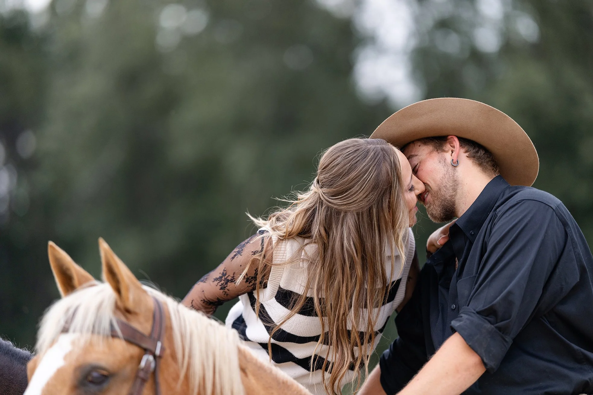 Amber leans down from her horse to kiss Isaac during their equestrian engagement photos in Poynette, Wisconsin, showcasing a tender moment surrounded by their horses and a wooded backdrop.