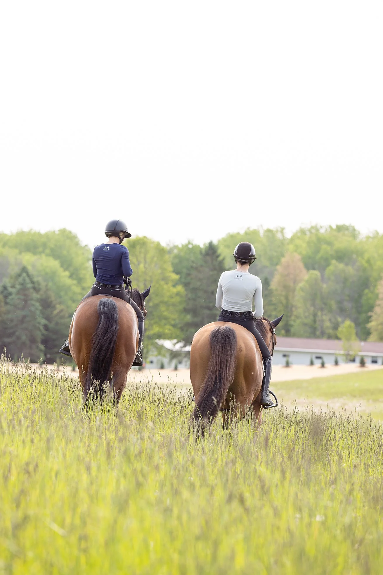 Two riders wearing STS Equestrian shirts trail riding through tall grass, viewed from behind.